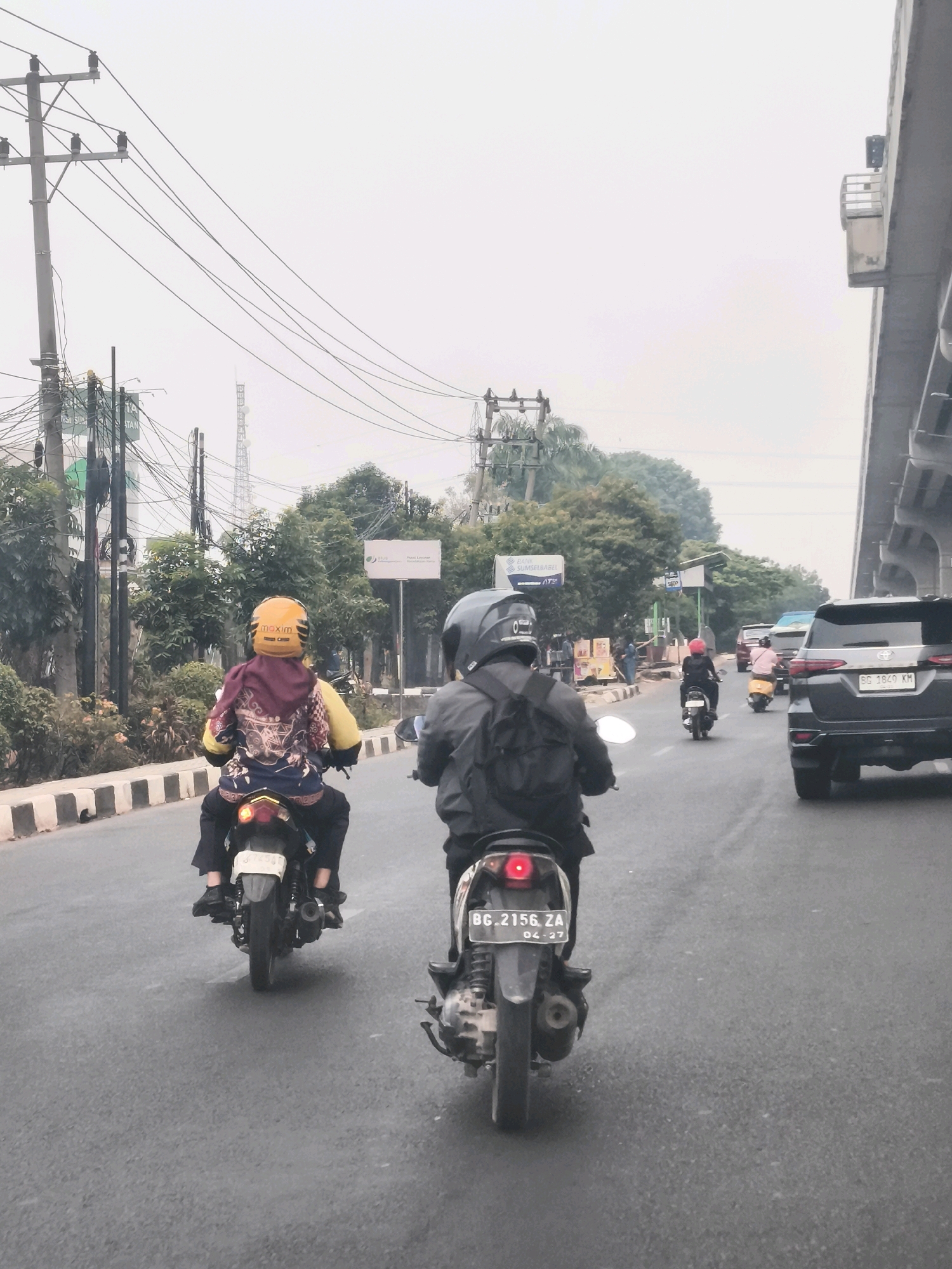 The image depicts a street scene in Palembang, Indonesia, characterized by hazy skies and dense traffic. In the foreground, two individuals are riding motorcycles. The motorcycle on the left is being ridden by a person wearing a yellow helmet with "maxim" written on it and a hooded jacket with intricate patterns. This rider is positioned closer to the viewer and is on the left side of the road, following a black bordered divider. The motorcycle on the right is ridden by a person wearing a gray helmet, with their back to the camera.
The middle ground reveals a black SUV with a license plate reading "BG 1849 KM," indicating it is registered in South Sumatra, where Palembang is located. Behind the SUV, several other motorcycles are visible, some carrying multiple passengers. The roadside is lined with trees and vegetation, with utility poles and power lines forming a prominent overhead structure, characteristic of urban infrastructure in Indonesia. In the distance, a transmission tower is visible against the overcast sky. The overall environment suggests a busy urban setting with moderate air pollution. A section of an elevated structure, possibly a bridge or overpass, is present on the right side of the frame. The road surface is asphalt.