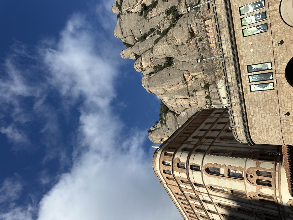 The image captures a panoramic view in Collbató, Spain, featuring mountainous terrain and architectural structures under a blue sky with scattered white clouds. The upper right and background are dominated by the imposing, light grey and tan conglomerate rock formations of the Montserrat mountains, exhibiting characteristic rounded peaks and steep faces, with sparse green vegetation clinging to crevices. In the mid-ground left, a multi-story building with a rounded corner is visible, constructed with alternating cream-colored plaster sections and reddish-brown brickwork. It features multiple arched windows on each of its five visible floors, and a white cylindrical object is atop its roof. In the lower right foreground, a stone-block structure with a curved facade presents several large, vertically oriented windows containing graphic banners that partially spell "MUS" and "EU" from "MUSEU". Three dark-colored streetlights are mounted along the upper edge of this foreground structure. Further up the mountainside, behind the foreground structure, smaller, light-colored stone buildings with arched windows are discernible on a terrace. No individuals or active subjects are present. Direct sunlight illuminates the scene, casting shadows on the left side of the prominent multi-story building.