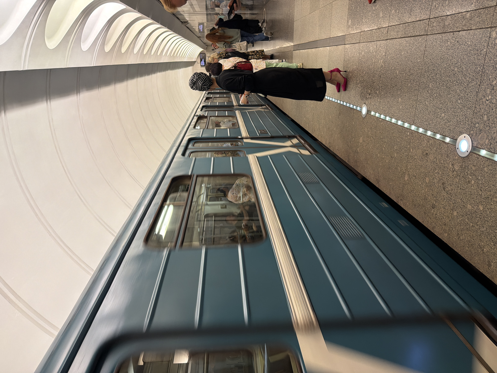 The image captures a subway station in Moscow, Russia. A teal-colored subway train is moving past the platform, creating a blur of motion. On the platform, several people are waiting. In the foreground, a woman dressed in black with a black and white patterned headscarf and bright pink heels stands looking at the train. Behind her, other individuals are also waiting, including a woman in a floral dress and another in a green hoodie. The station features a high, arched ceiling with circular light fixtures and a tiled wall. A strip of lights runs along the edge of the platform.