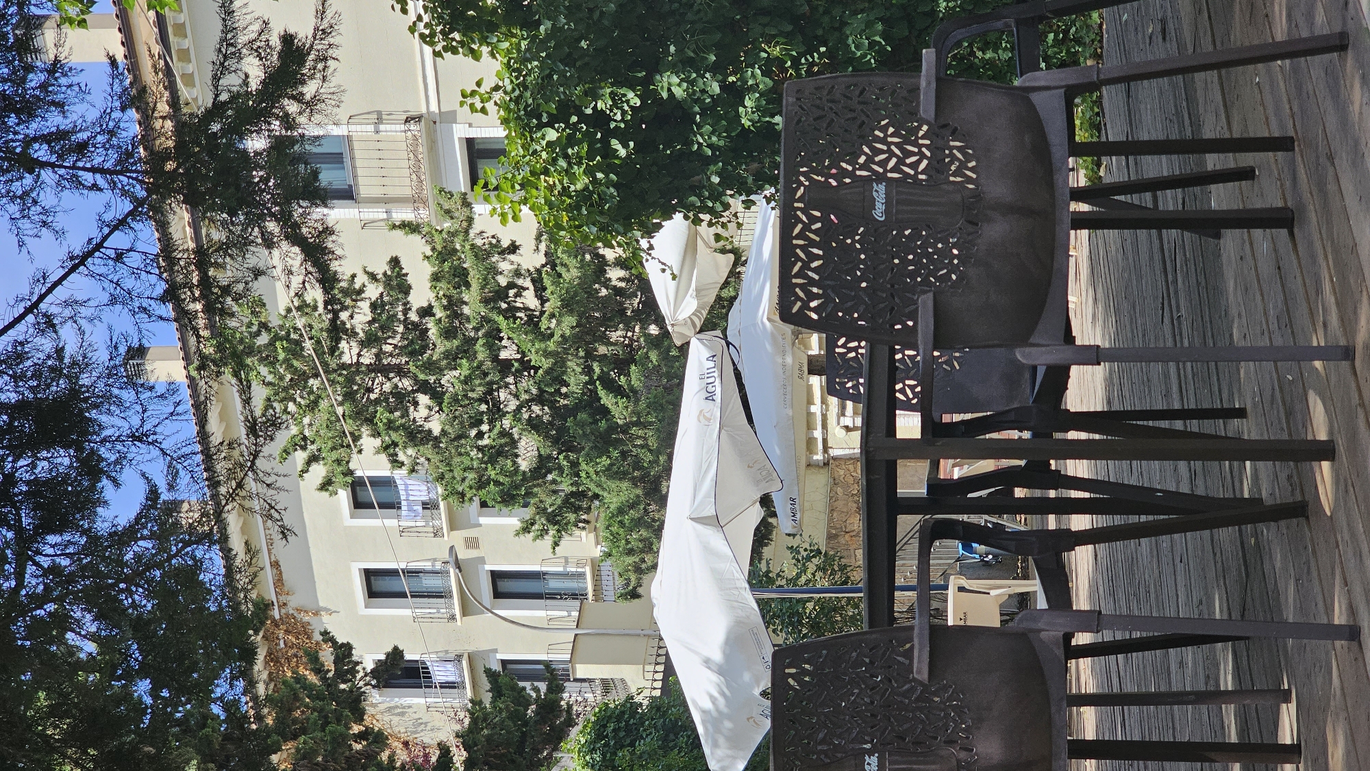 This media file depicts an outdoor seating area with a building in the background. The scene is set on a wooden deck, with several dark metal chairs and tables, some with ornate, patterned backs. The chairs are arranged around the tables, suggesting a space for dining or relaxation.
In the background, a light-colored building with multiple floors and windows is visible. Some windows have wrought-iron balconies. Lush green trees and foliage surround the building and extend into the foreground, creating a natural, outdoor ambiance.
Several white umbrellas are present, some partially furled or open, suggesting that this is a cafe, restaurant, or outdoor bar. One umbrella prominently displays the text "EL ÁGUILA" in black lettering, along with other smaller, less legible text, possibly branding or advertising. Another umbrella shows the text "COCA-COLA".
The lighting suggests it is daytime, with sunlight casting shadows. The weather appears to be pleasant, conducive to outdoor activities. The overall atmosphere is relaxed and inviting, typical of a Mediterranean setting. The location context provided is Alhama De Aragón, Spain, which aligns with the visual cues of a building with balconies and abundant greenery. There are no people visible in the scene, but the setup implies a place where people would gather.