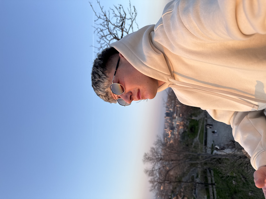 The image displays a young male, positioned from the chest up, gazing towards the right, with the city of Plovdiv, Bulgaria, visible in the background from an elevated vantage point, likely one of its hills. He wears a light beige hooded sweatshirt with visible drawstrings and aviator-style sunglasses, which reflect orange and pink light from the setting sun. His hair is short, faded on the sides, and lighter on top. The sky above is clear and blue, transitioning to warmer hues near the horizon. Below, a sprawling cityscape extends into the distance, partially obscured by barren, deciduous trees and ancient-looking stone structures on the hillside. A paved pathway with several indistinct pedestrian figures is visible further down the slope. The scene is illuminated by the warm, directional light of a late afternoon or early evening sun.