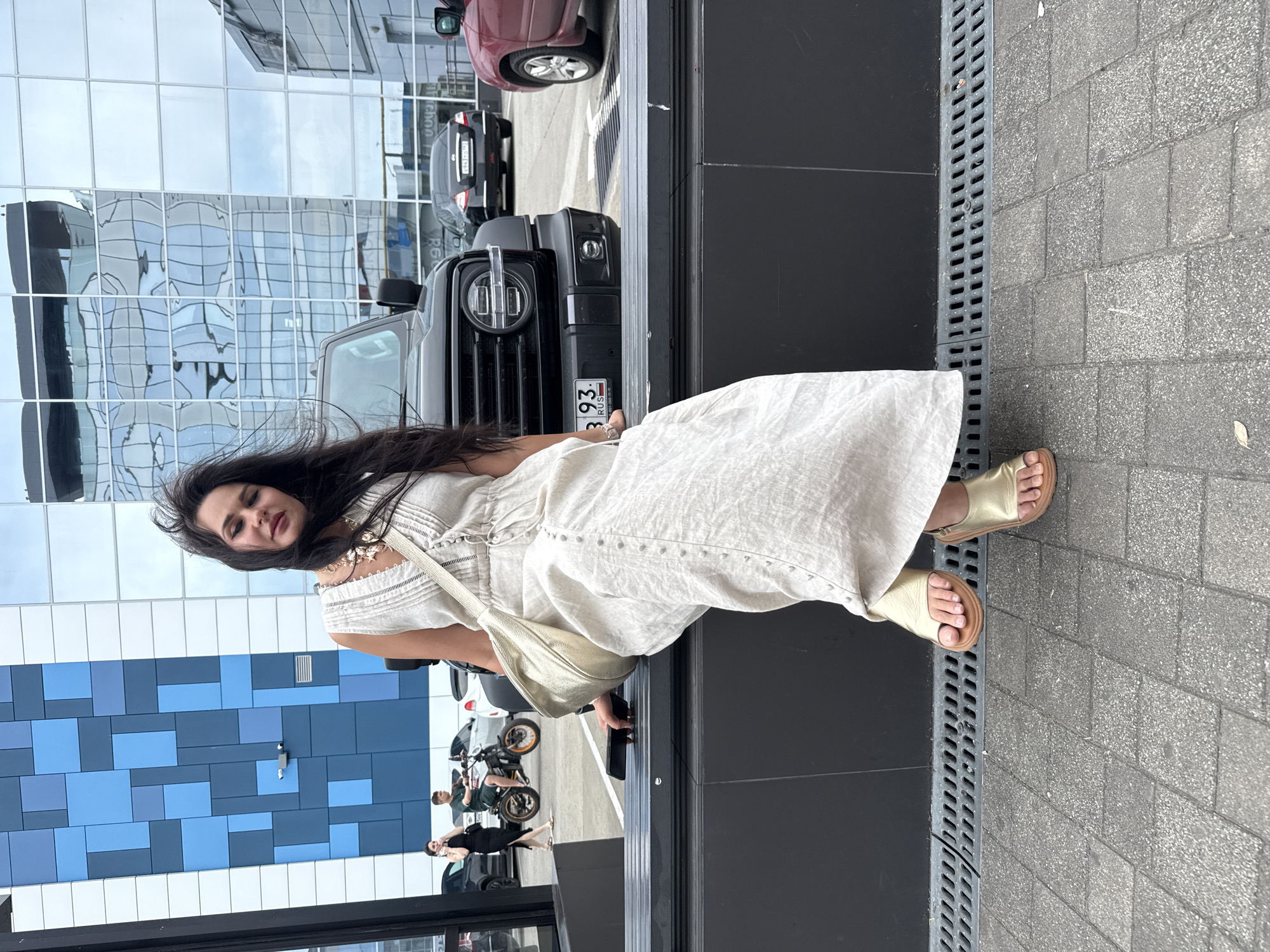A woman with long, dark hair is sitting on a dark bench in front of a black SUV. She is wearing a long, light-colored linen dress and gold sandals. She is holding a black smartphone in her right hand. The background features a building with a modern glass facade that reflects the sky and surrounding structures. To the left, a blue and white patterned wall is visible. In the middle ground, a woman in a black dress is talking on her phone, and a man is riding a motorcycle. The pavement is made of gray interlocking bricks. This scene is located in Sochi, Russia, likely in a parking lot or public area adjacent to a modern building.