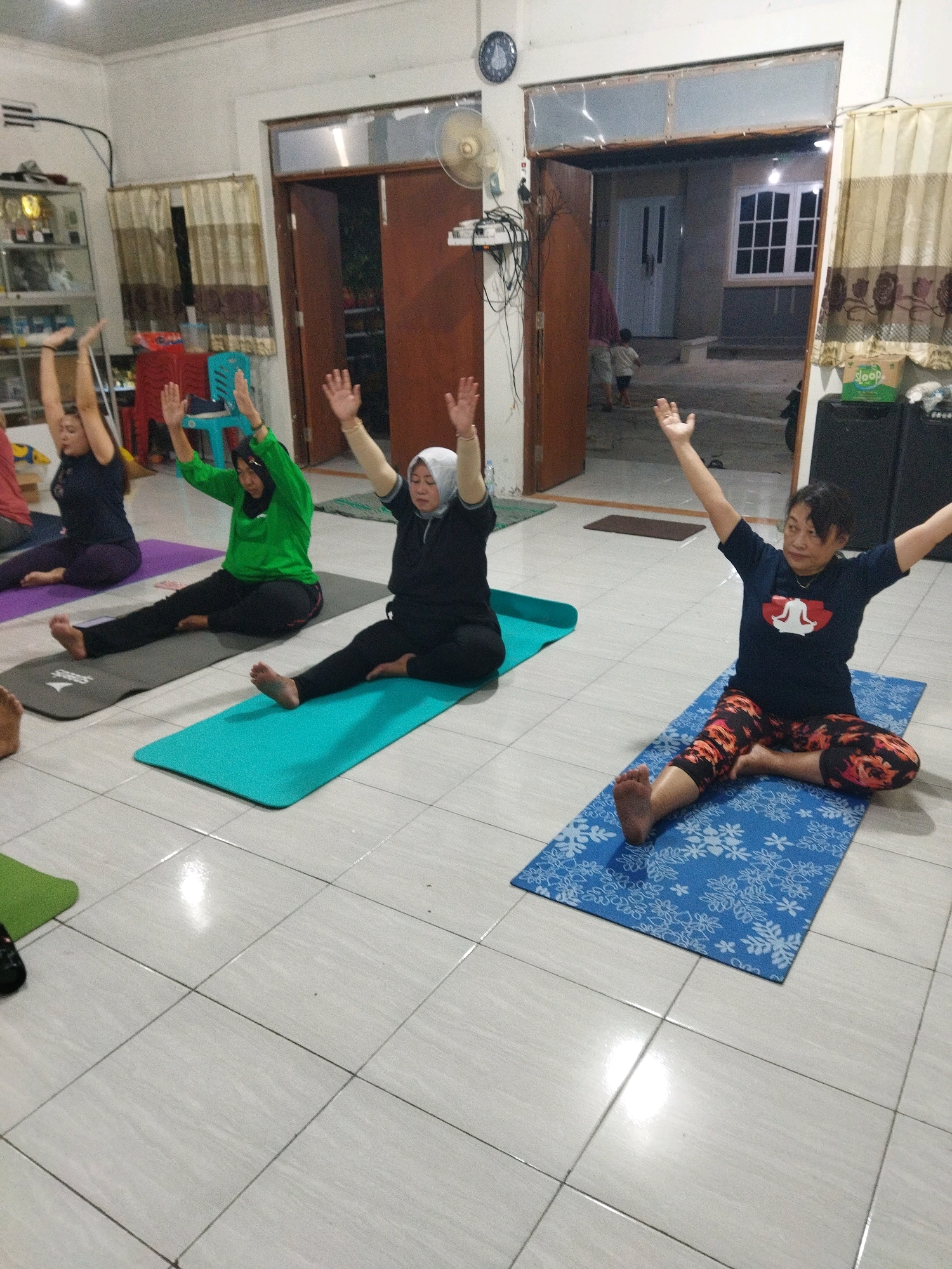 The image depicts an indoor scene, likely a community space or a home, with four women practicing yoga on yoga mats. The floor is tiled with a light grey pattern. The women are positioned from left to right, each on their own mat. The woman on the far left is wearing a dark t-shirt and purple leggings. Next to her, a woman in a bright green long-sleeved shirt and black leggings is doing the same pose. In the middle, a woman in a black outfit and a white hijab is also in a yoga pose. On the far right, a woman in a dark blue t-shirt with a white graphic and patterned leggings is performing the yoga pose. All women have their arms raised upwards. In the background, there is an open doorway leading to an outdoor area with a house visible. There are also curtains, a shelf unit with items, and a fan. The lighting suggests it is daytime, possibly late afternoon, with artificial lights also in use. The presence of a fan and the style of the building visible through the doorway hint at a tropical climate. This scene could be located in Semarang, Indonesia, given the common practice of yoga in community settings and the architectural elements.