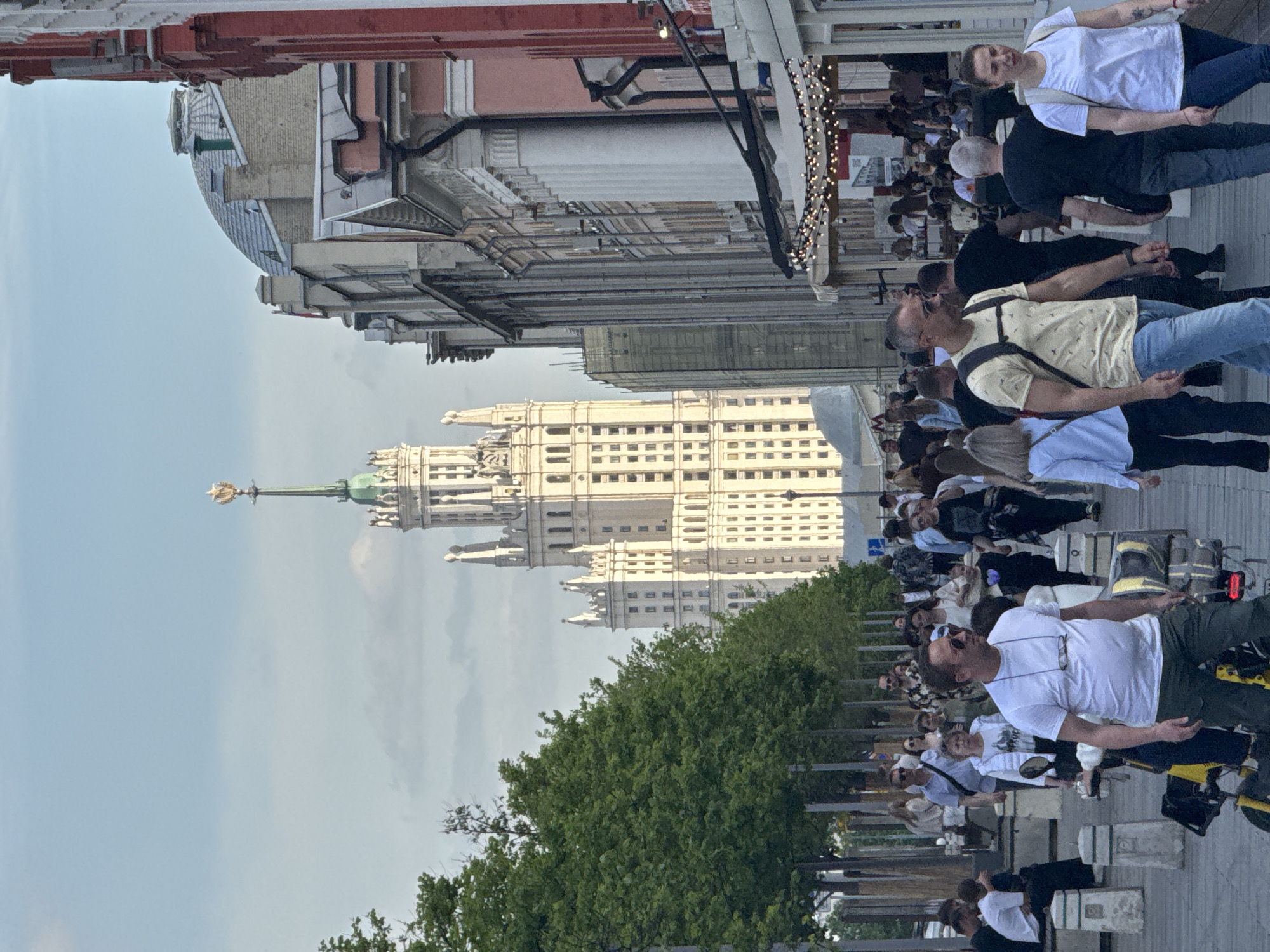 The image shows a street scene in Moscow, Russia, with the Kotelnicheskaya Embankment Building, one of the Seven Sisters skyscrapers, visible in the background. A crowd of people is walking along the sidewalk, with some riding scooters. Buildings line the street, and trees are visible in the foreground. The sky is overcast.
