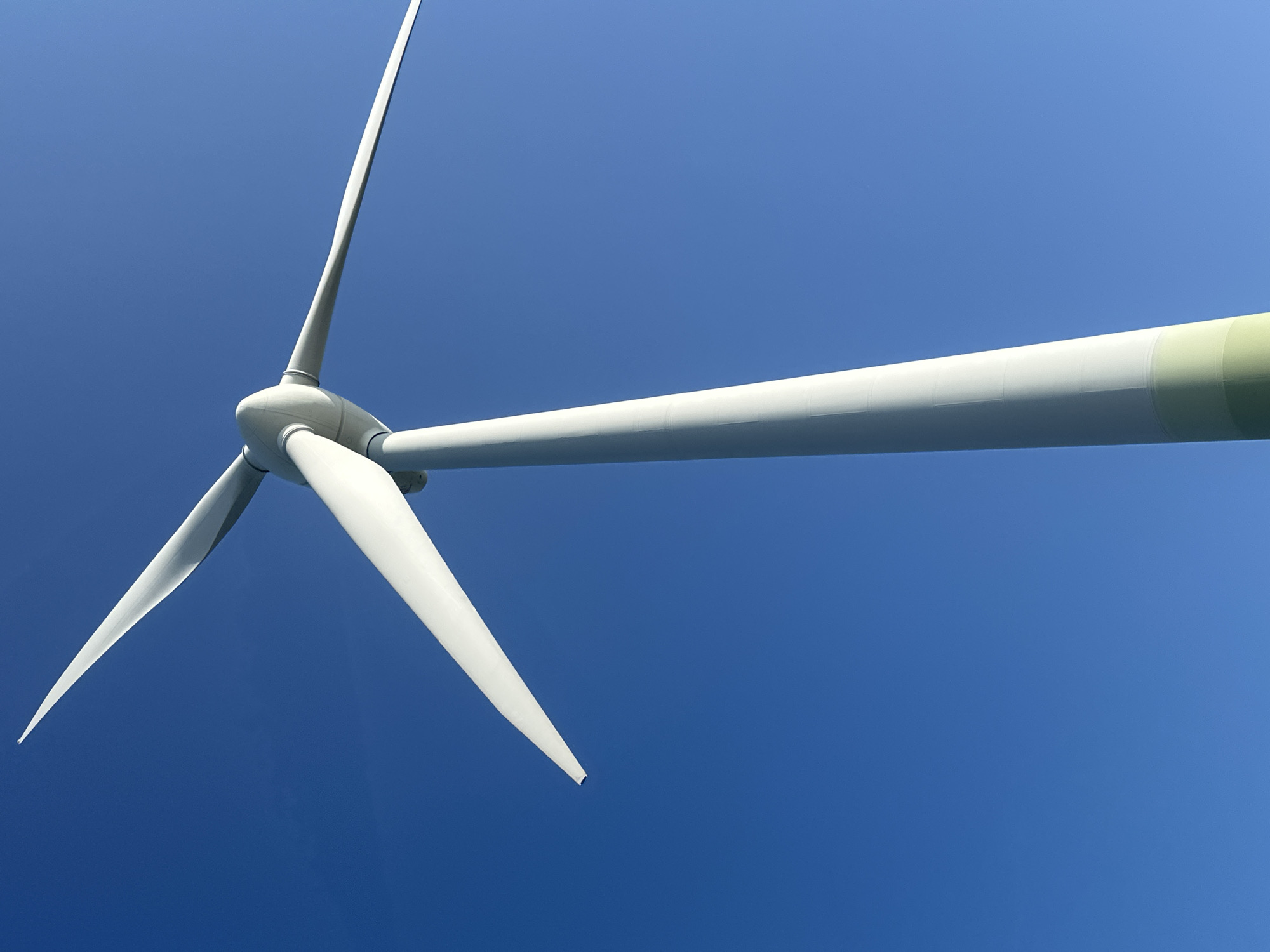 A low-angle, full-shot photograph captures a wind turbine against a vibrant, cloudless blue sky. The turbine's large, white blades are extended, with one blade positioned prominently in the foreground, pointing towards the upper left. The central mast of the turbine is also visible, extending upwards from the bottom of the frame and tapering slightly as it rises. The lower portion of the mast displays a distinct section of light green paint, differentiating it from the white upper section. The image composition emphasizes the scale and dominance of the wind turbine against the vast expanse of the sky, suggesting an open, possibly rural or coastal environment. There are no people or animals present in the frame. The scene is static and still, with no discernible action occurring. The image's clear focus and bright lighting indicate it was taken on a sunny day. Given the question's request for location, it is reasonable to infer this photograph could be of a wind turbine located in the vicinity of Liepaja, Latvia, a city known for its coastal geography and wind energy infrastructure.
