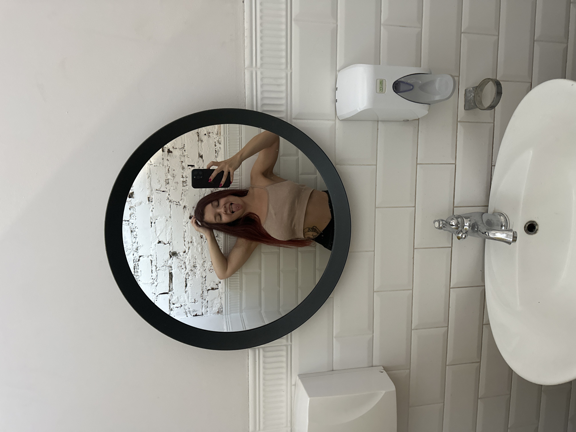 A young woman with long, red hair is taking a mirror selfie in a public restroom. She is wearing a beige crop top and black bottoms. She is sticking her tongue out and holding her phone in her right hand. Her left hand is resting on her head. The restroom features white tiled walls and a white sink with a chrome faucet. There is also a white soap dispenser mounted on the wall. The background shows a white brick wall. The scene appears to be in a modern establishment. The image does not provide any specific clues or indicators to determine the city of Vladimir, Russia.