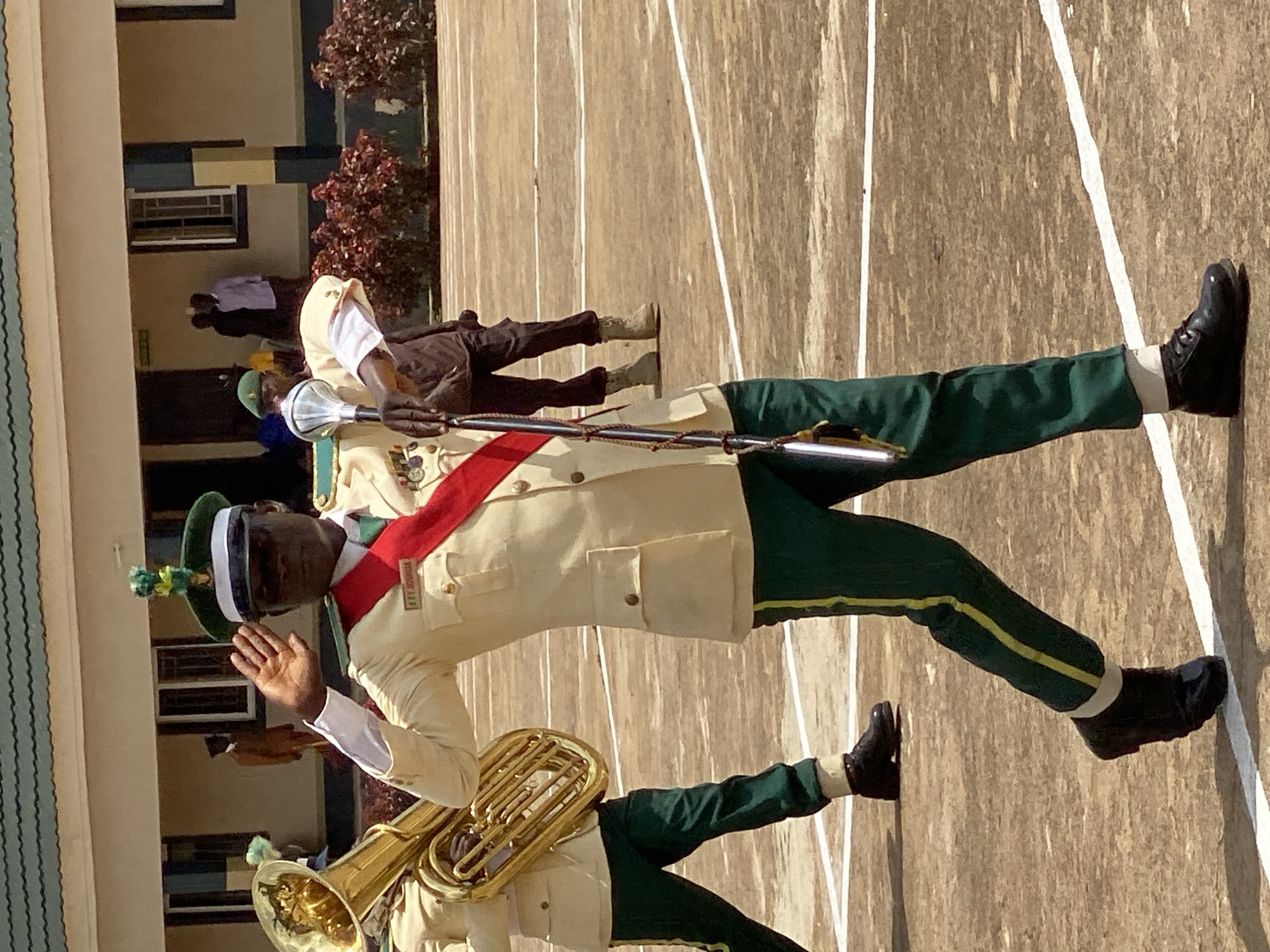The image depicts a group of people in uniform marching outdoors, likely in Kaduna, Nigeria. The primary subject is a young man in the foreground, wearing a cream-colored uniform jacket with epaulets and a red sash across his chest. He has a dark green cap with a white band and a decorative pom-pom. His right hand is raised in a greeting gesture, and he is wearing a white shirt with dark green trousers that have a yellow stripe down the side. He is holding a ceremonial baton. Behind him, another person in a similar uniform can be seen with a brass instrument, possibly a tuba. Further back, more individuals in uniform are visible, some holding batons and others appearing to be part of a marching band. They are on a paved surface marked with white lines, suggesting a parade ground or sports field. In the background, there is a building with windows and some trees with red foliage. The lighting suggests it is daytime, possibly late morning or afternoon, with clear weather. The uniforms and the presence of musical instruments indicate a formal event or procession, possibly a school parade or a cultural celebration. The overall scene conveys a sense of order and discipline. Visible text on the sash reads "ROYAL SCOUTS".
