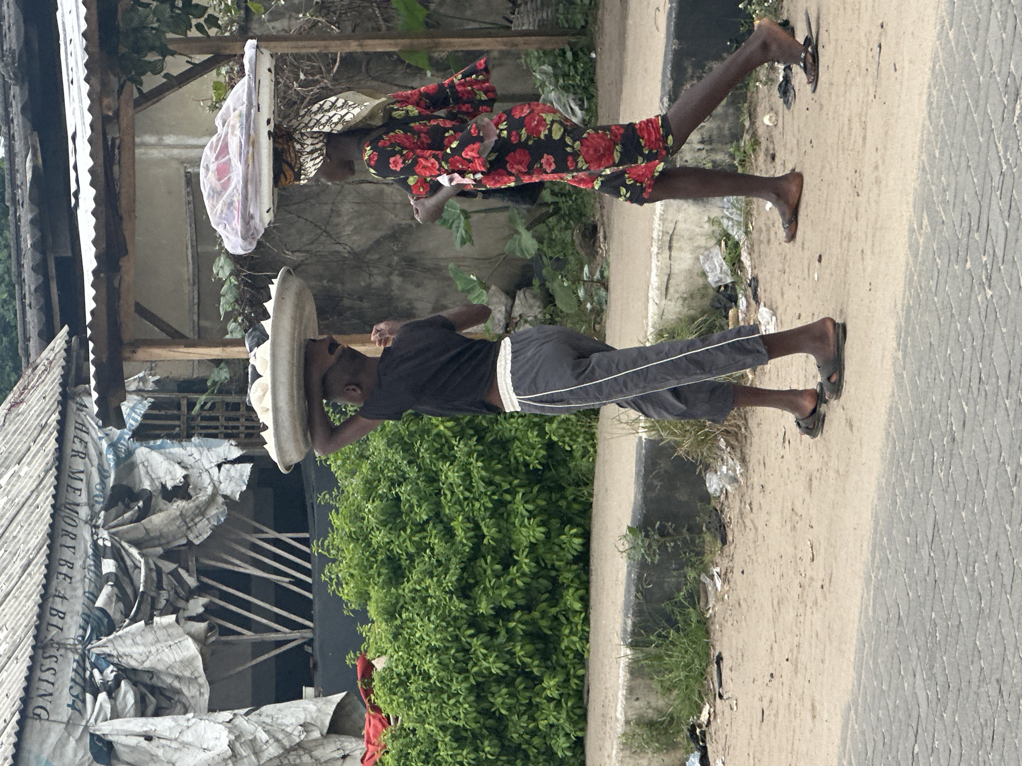 In Badagry, Nigeria, two individuals are walking on a dirt road beside a concrete barrier. The person on the left, a male, is carrying a large white bowl filled with round, light-colored food items on their head. They are wearing a black t-shirt and grey sweatpants with white stripes. The person on the right, a female, is wearing a black dress with a red floral pattern and a patterned headscarf. She is also carrying a white tray on her head, covered with a transparent plastic sheet that encloses various colorful items, possibly food or merchandise. Behind them, there is a dense green bush, and further back, a dilapidated structure with a corrugated metal roof and tarpaulin coverings, suggesting a market stall or informal shelter. The ground is sandy with scattered debris.