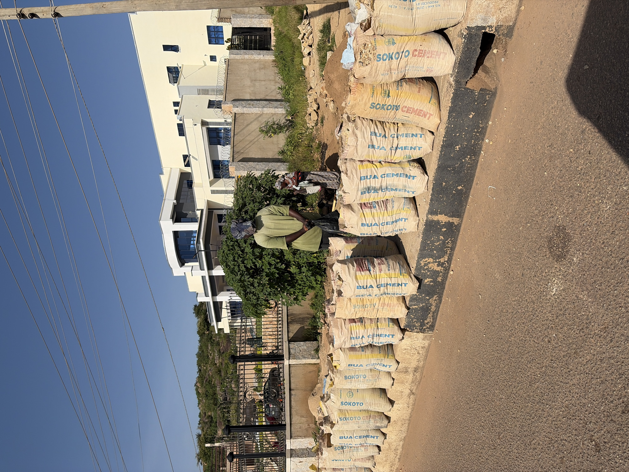 In Gura Top, Nigeria, a street scene unfolds with a row of cement bags lining a curb. Two individuals are present, with one person, a man wearing a light green t-shirt and a head covering, standing on the left side of the image, holding a black plastic bag. To his right, a woman in a patterned dress stands next to a pile of dirt and rocks. Behind them, a modern, multi-story building with cream-colored walls and blue-tinted windows stands prominently. The area is fenced with a decorative black metal fence and barbed wire. The sky is clear blue, and utility wires are strung across the frame. The road surface in the foreground is asphalt.