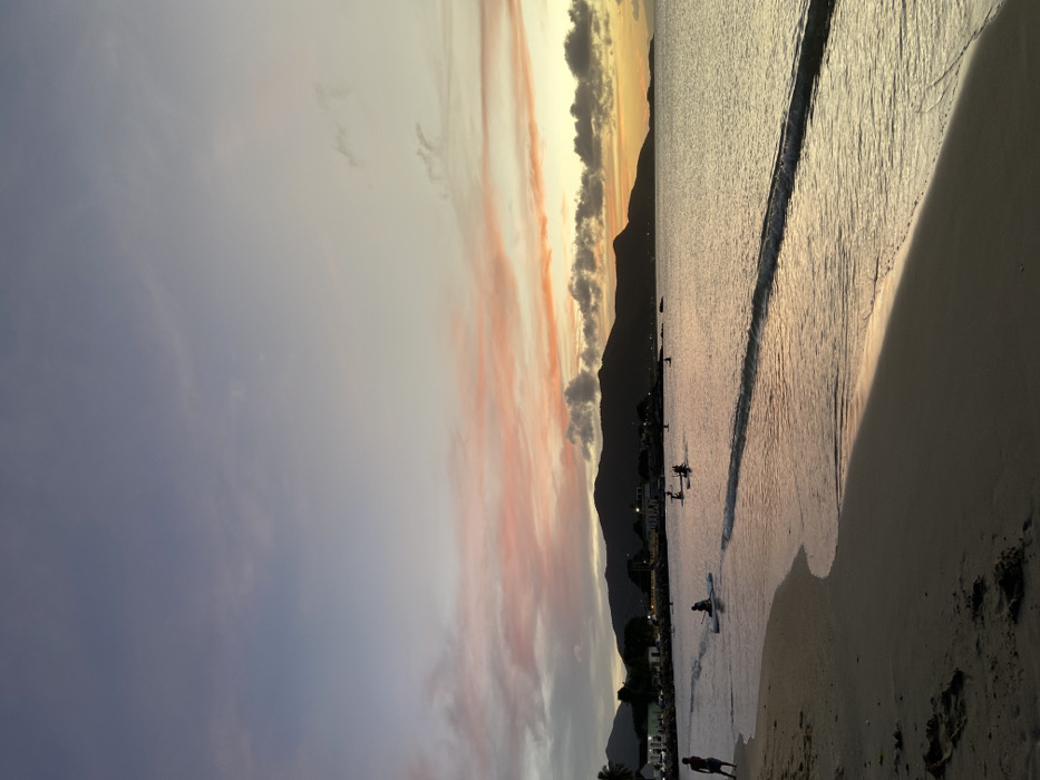 A beach scene during sunset. Soft waves roll onto the sand, reflecting the golden light. Silhouetted mountains form a backdrop. People are seen on paddleboards in the water and walking along the shoreline. The sky is a mix of blues, grays, pinks, and yellows. Buildings and trees line the base of the mountains.