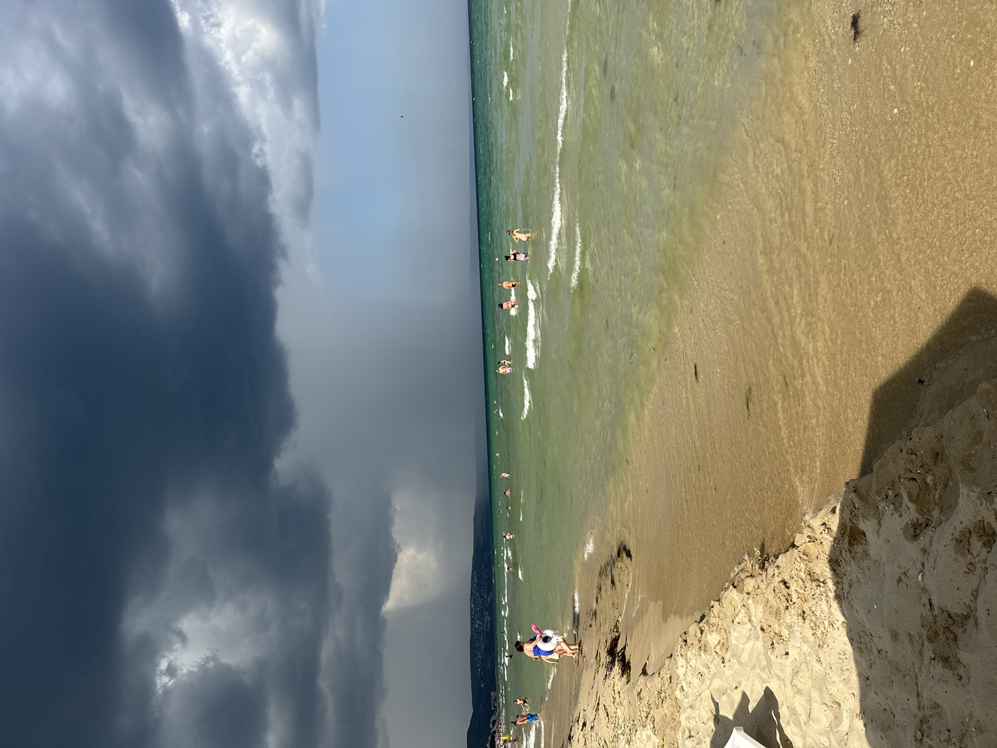 The image depicts a beach scene in Osenovo, Bulgaria, under a dramatic sky. Dark, ominous storm clouds dominate the upper half of the frame, with shafts of rain visible in the distance. The sea is a clear, greenish hue, with moderate waves breaking near the shore.
Several people are present on the beach and in the water. On the sandy shore in the foreground, a woman wearing a blue swimsuit and a girl with a yellow float are walking towards the water. Further down the beach, other individuals are visible. In the water, a group of people, primarily women, are scattered across the sea, some wading, others swimming.
The beach itself is sandy, with some dried seaweed and debris. The shoreline curves gently, meeting the water. In the background, a distant coastline with greenery and buildings can be seen. The overall atmosphere is one of contrast, with the impending storm creating a sense of urgency against the backdrop of people enjoying a day at the sea.