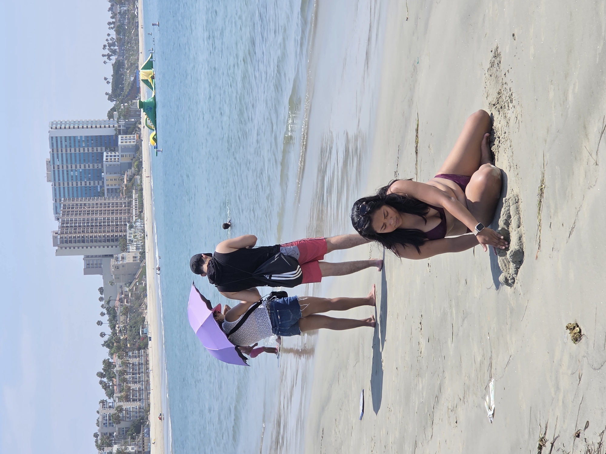 The image shows a beach scene in Long Beach, California. In the foreground, a woman wearing a burgundy bikini is sitting on the sand and building a sandcastle. Behind her, a couple stands on the wet sand near the water's edge. The woman is holding a purple umbrella, and the man is wearing a black tank top and red shorts. In the background, the ocean stretches out to the horizon with a few people swimming. Beyond the beach, there are buildings and palm trees lining the coast. An inflatable water park is visible in the ocean. The sky is clear and blue, indicating a sunny day.