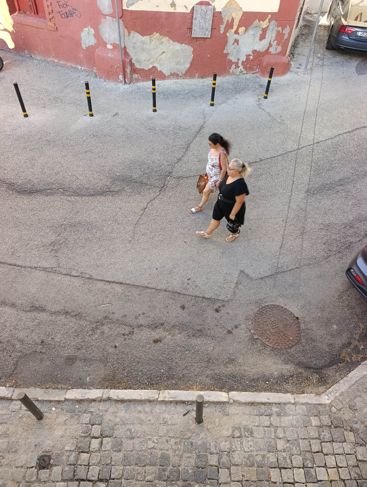 The image shows a street scene in Setúbal, Portugal, as viewed from above. In the middle ground, two women are walking on a paved street. The woman on the left is wearing a floral print dress and carrying a brown bag. The woman on the right is wearing a black dress and carrying a black bag. They are walking away from the viewer.
In the background, a red building with peeling paint and graffiti is visible. Several bollards with yellow and black stripes are placed along the edge of the street. A dark-colored car is parked on the right side of the street, and a manhole cover is present on the asphalt.
The foreground shows a cobblestone sidewalk with a metal bollard. The overall environment appears to be an urban street with signs of wear and tear.