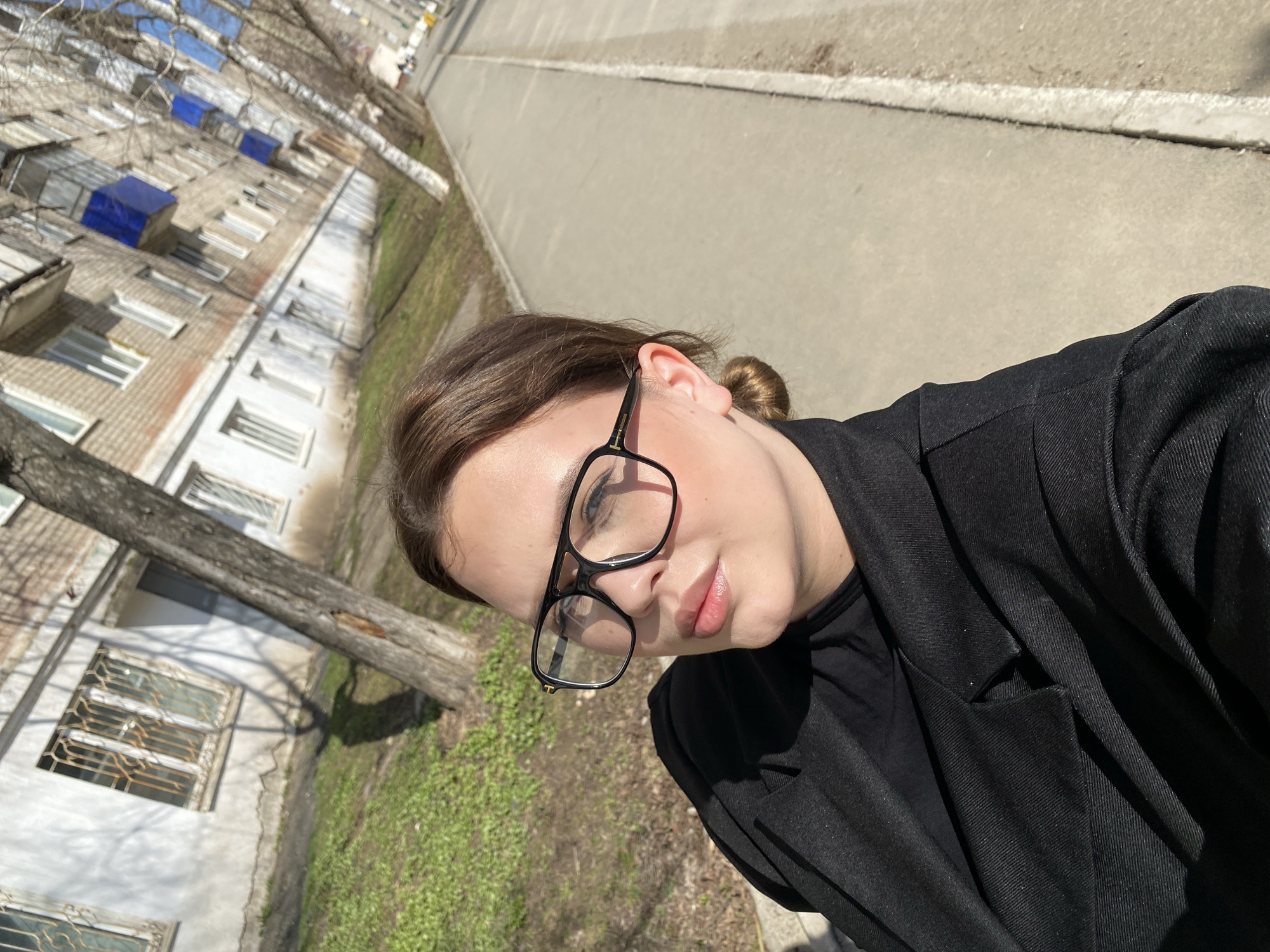 A selfie-style image, significantly tilted clockwise, features a young woman outdoors in what appears to be Chernushka, Russia. The woman, the primary subject, has light brown hair styled in a bun and wears black-framed glasses, a black top, and a black jacket. She is positioned in the foreground, looking towards the camera with a neutral expression, her lips slightly parted.
The background reveals an urban environment under bright daylight. To the left, a multi-story residential building with numerous windows, some fitted with metal grates, stretches into the distance. Several distinctive blue covers or structures are visible on the building's roofline or upper levels. A bare tree trunk runs horizontally across the midground, suggesting late winter or early spring. The ground consists of a dirt path with patches of green grass, leading to a wider paved road or large sidewalk with a visible curb. The bright sunlight casts clear shadows, indicating a sunny day. No text is visible in the image.