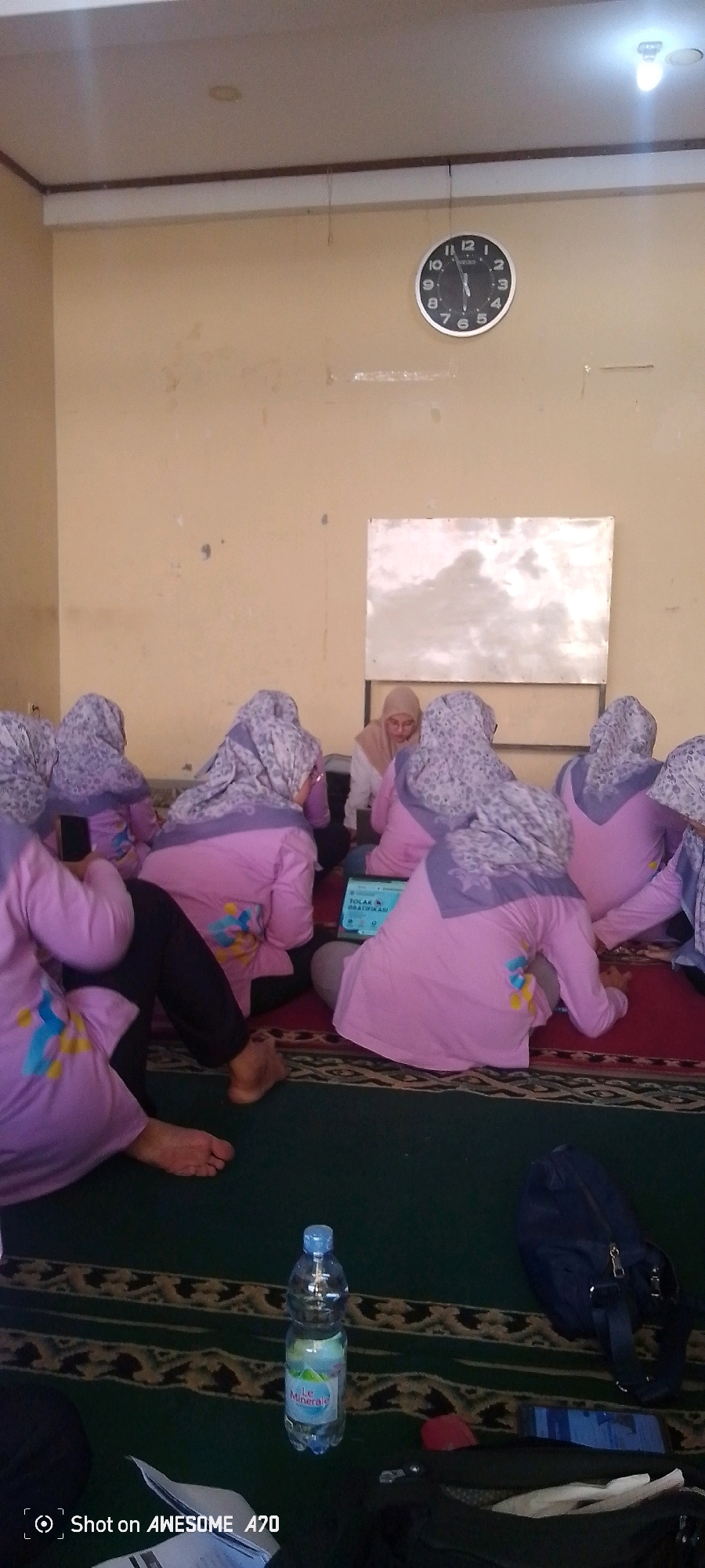 A group of women wearing purple shirts and headscarves are sitting on a carpeted floor in a room. One woman in a beige headscarf is facing a laptop. There is a whiteboard on the wall behind them. A bottle of water and a blue bag are on the floor in the foreground. The room appears to be a classroom or meeting space, possibly in the city of Bandung, Indonesia, given the presence of Indonesian text on the laptop screen. The image is a photograph, and the lighting suggests it was taken indoors during the day.