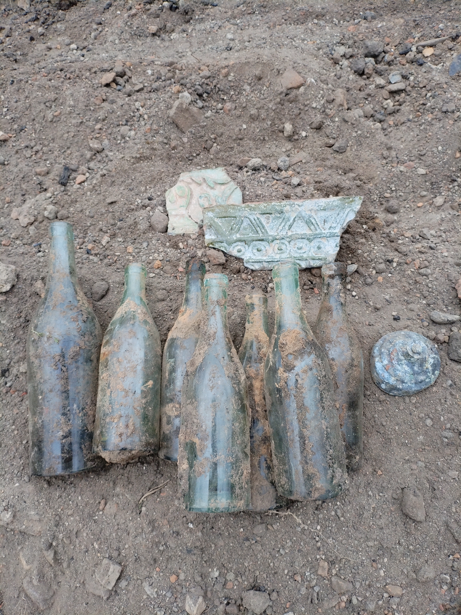 In Bialynichy, Belarus, an archaeological dig has unearthed a collection of glass bottles and ceramic fragments. Seven glass bottles, some partially filled with dirt, are arranged in a group on the ground. They appear to be old, with varying shades of green and a layer of dust and soil adhering to their surfaces. To the right of the bottles, a circular object with a textured surface, possibly a lid or a large button, is visible. Behind the bottles, several pieces of a light-colored, possibly ceramic, object are scattered. One prominent piece features a decorative embossed pattern of triangles and circles. The objects are situated on loose soil mixed with small stones and debris, indicative of an excavation site. There are no individuals present in the image.