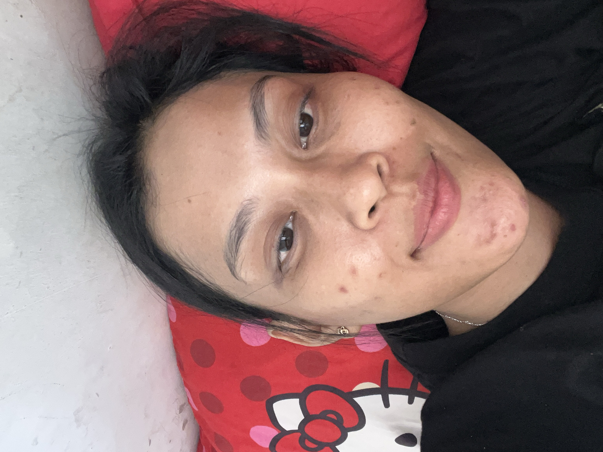 A selfie taken by a person lying on a red pillow with a Hello Kitty design. The person has dark hair, brown eyes, and fair skin with visible blemishes. They are wearing a black shirt. The background appears to be a wall. The image is a close-up shot of the person's face and upper body. The location is not discernible from the image itself.