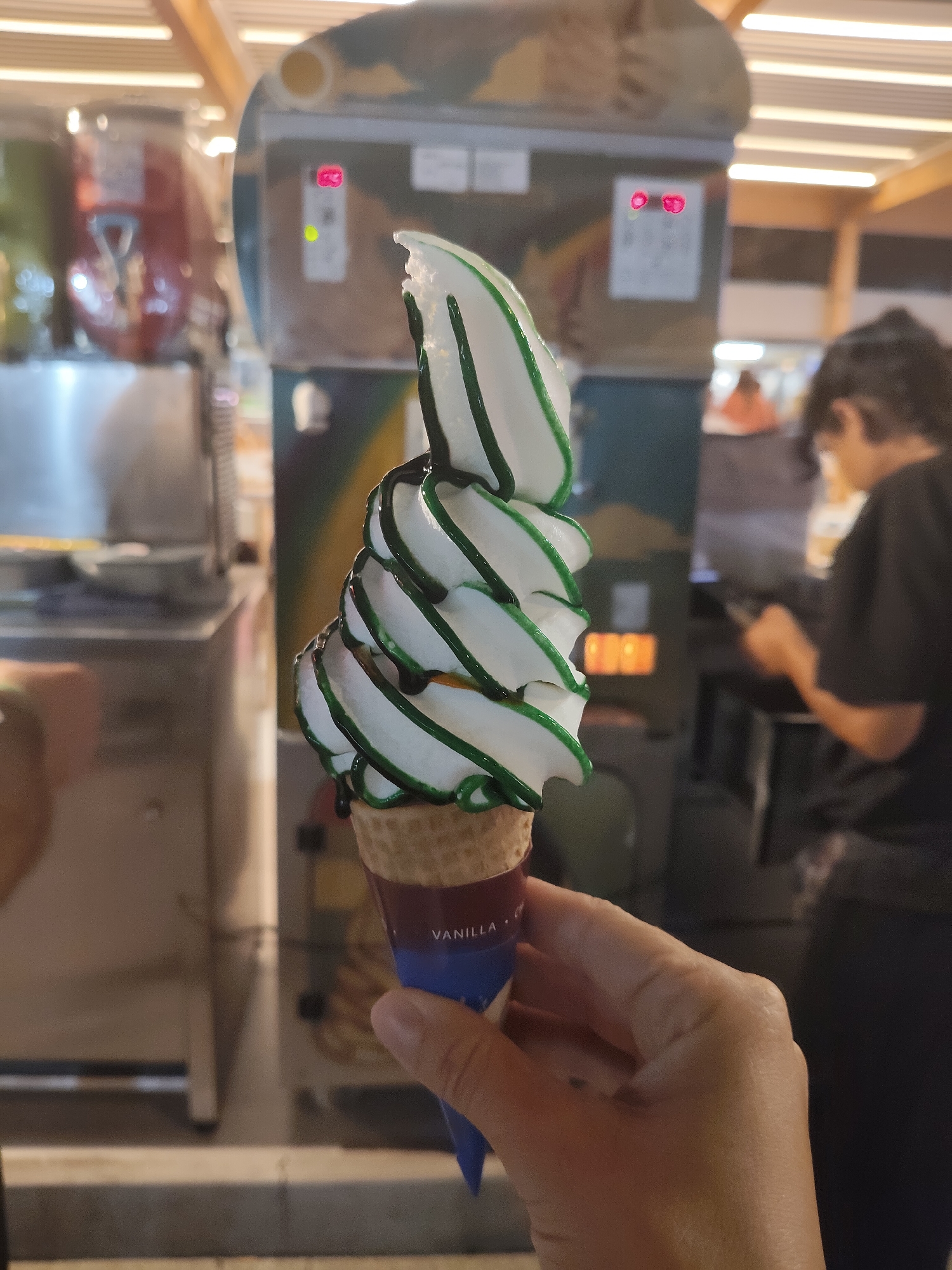 The image shows a close-up of a vanilla soft-serve ice cream cone with green syrup drizzle held by a person's hand. The ice cream is swirled and topped with the green syrup. In the background, a soft-serve ice cream machine is visible, along with what appears to be a beverage dispenser. A person with dark hair, wearing a dark shirt, is blurred in the right background, seemingly interacting with a device. The environment suggests an indoor establishment, possibly a food court or a kiosk within a mall or entertainment venue, with lighting fixtures visible on the ceiling. The setting is not specifically identifiable as Constanța, Romania, nor are there any clear landmarks or cultural indicators to suggest this location.