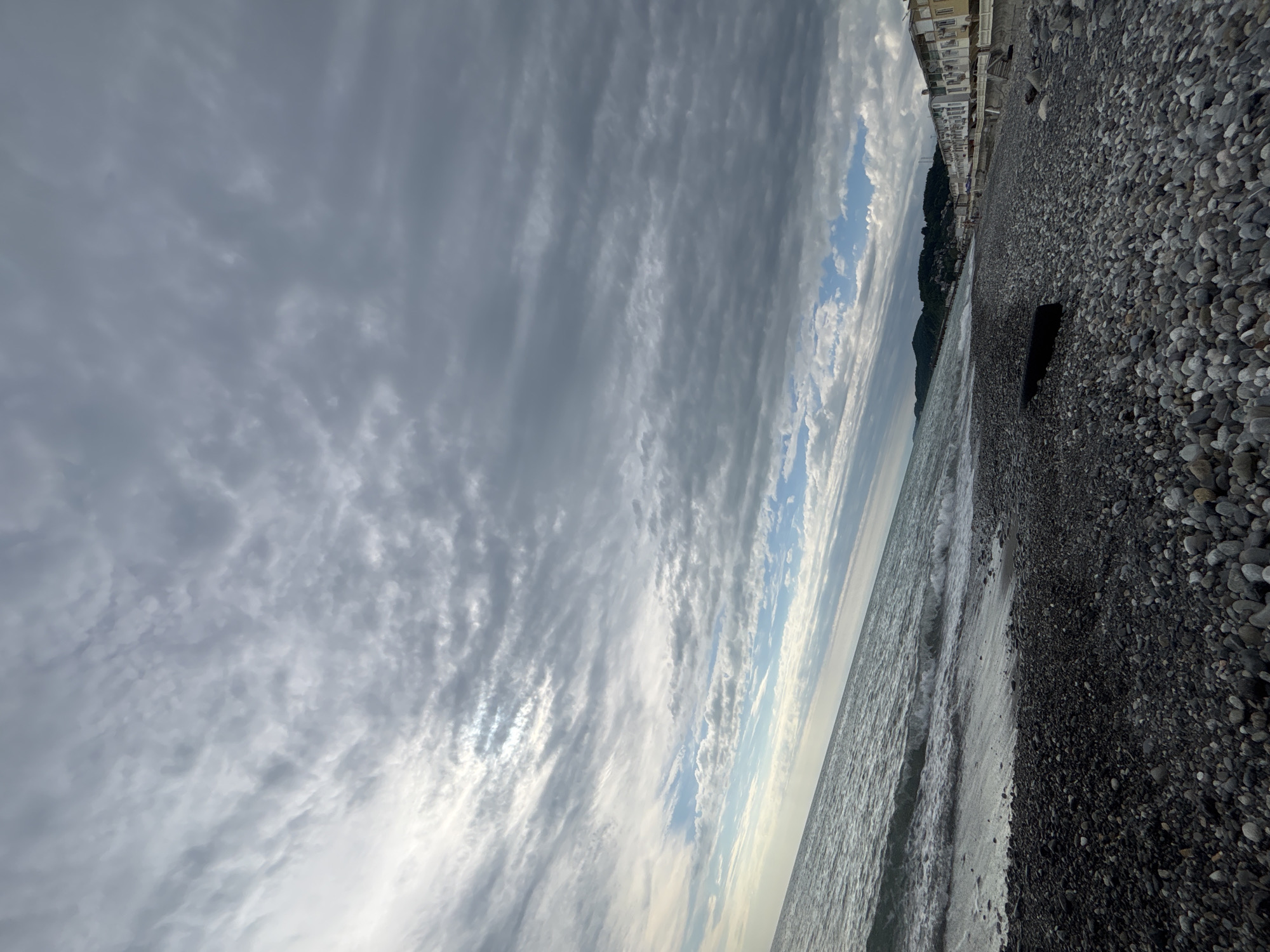 The image shows a beach in Sochi, Russia. The beach is composed of grey pebbles. The sea is choppy and grey. The sky is overcast with striated clouds. In the distance, there are buildings along the coastline. The image is taken at an angle.