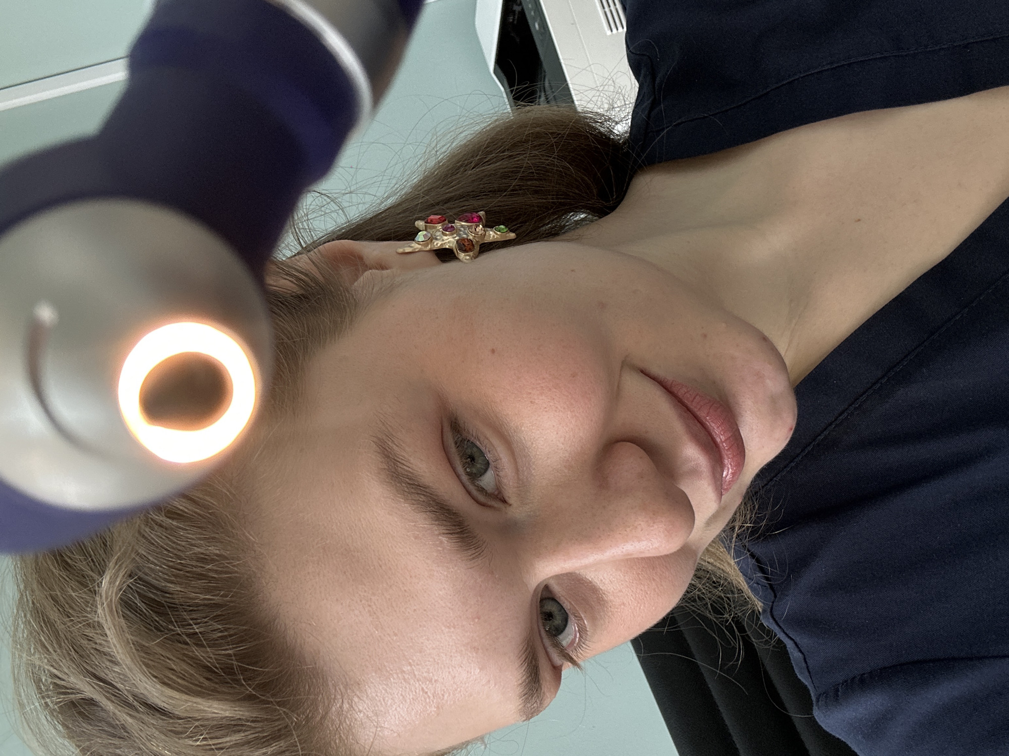 A fair-skinned woman is viewed from a low angle, looking directly at the viewer. A medical examination light is positioned close to her left ear, casting light on her face. She has fair hair and is wearing a colorful earring in her left ear. The woman is wearing a dark blue shirt. A portion of a medical device can be seen behind her head on the upper right part of the image. The image's background is nondescript, but suggests a clinical setting. There is no contextual information to identify the location as Kazan, Russia.