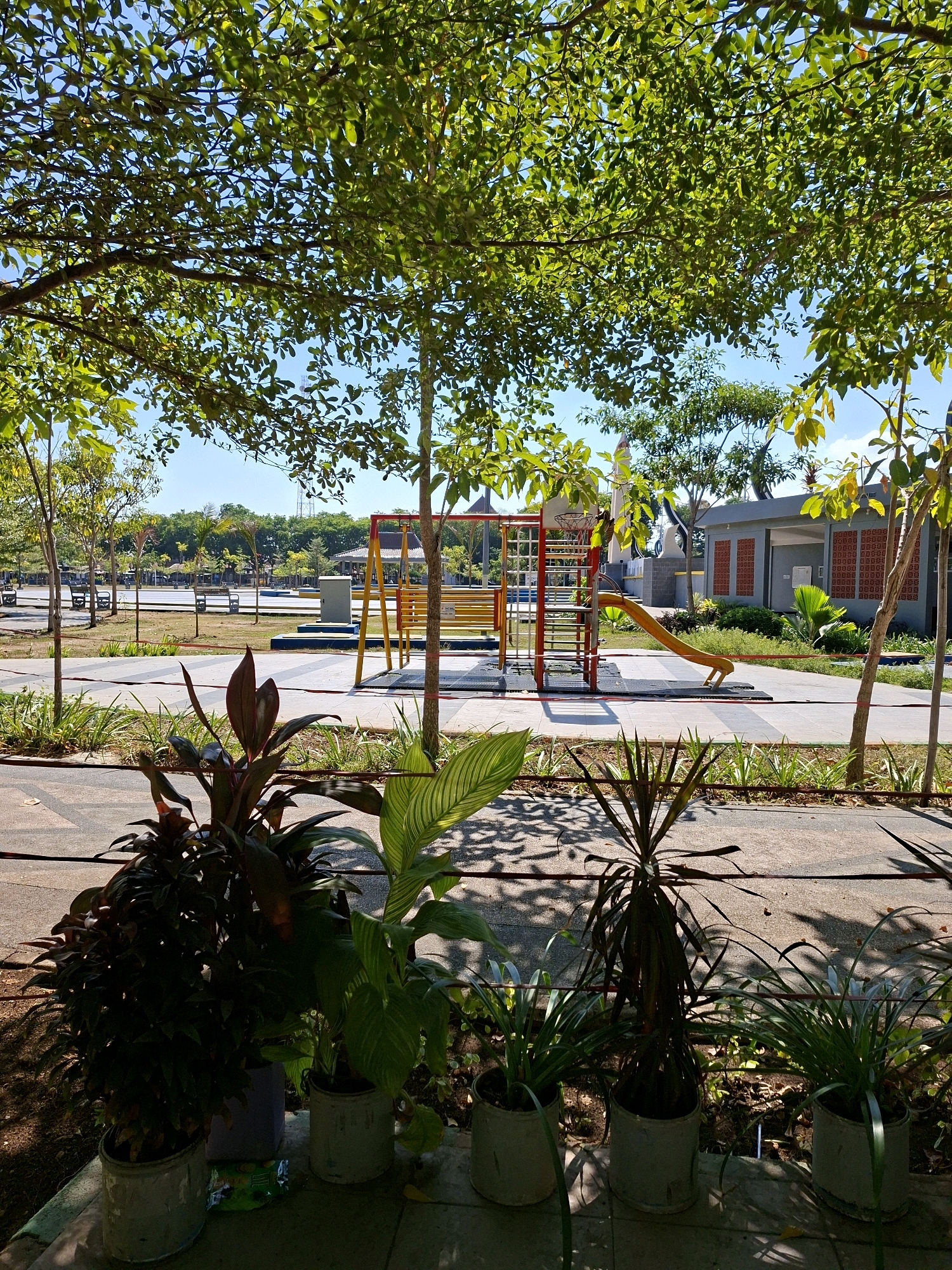 This is a sunny outdoor scene in Gunung Sekar, Indonesia. In the foreground, several potted plants are arranged, including dark red-leafed plants and plants with green and white striped leaves. The ground in the foreground is paved with textured concrete. Behind the plants, a walkway paved with gray and white tiles leads into the background. To the right of the walkway is a children's playground with a yellow slide and a red and yellow climbing structure. Further back, there are benches and trees. In the distance, a building with a gray facade and red patterned brick accents can be seen. The sky is clear and blue. The image captures a peaceful park setting on a bright day.