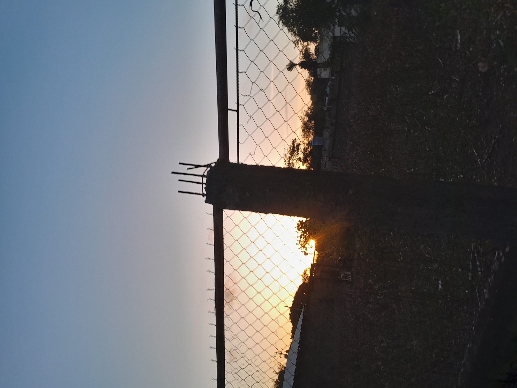 The image depicts a sunset viewed through a chain-link fence in Santa Ana, Venezuela. In the foreground, a concrete fence post with exposed rebar is visible, supporting the fence. Beyond the fence, the setting sun casts a warm glow across the sky, illuminating the chain-link mesh. Silhouetted against the bright sky are trees, some buildings with corrugated metal roofs, and utility poles. The ground in the foreground is dark and appears to be covered with vegetation or debris. The overall impression is a rural or semi-rural setting at dusk.