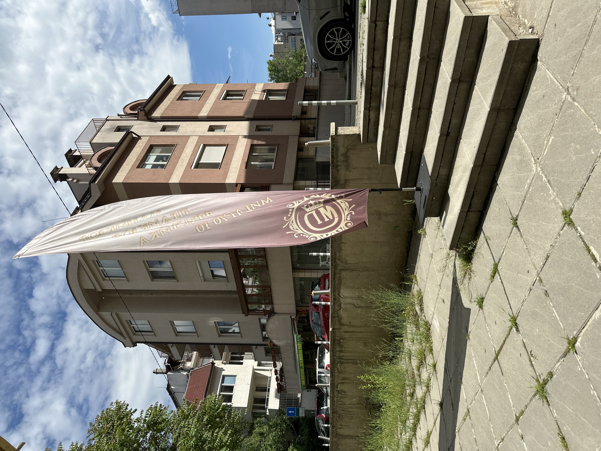 The image shows a street scene in Sofia, Bulgaria. A building with brown and beige exterior is visible, with a large banner in front displaying the logo "IM" and text in Bulgarian. Cars are parked along the street in front of the building. The photograph is taken at a low angle, looking up at the building and banner. Concrete stairs are visible on the right side of the image, with grass growing between the slabs. The sky is cloudy. The photo has been rotated 90 degrees clockwise.