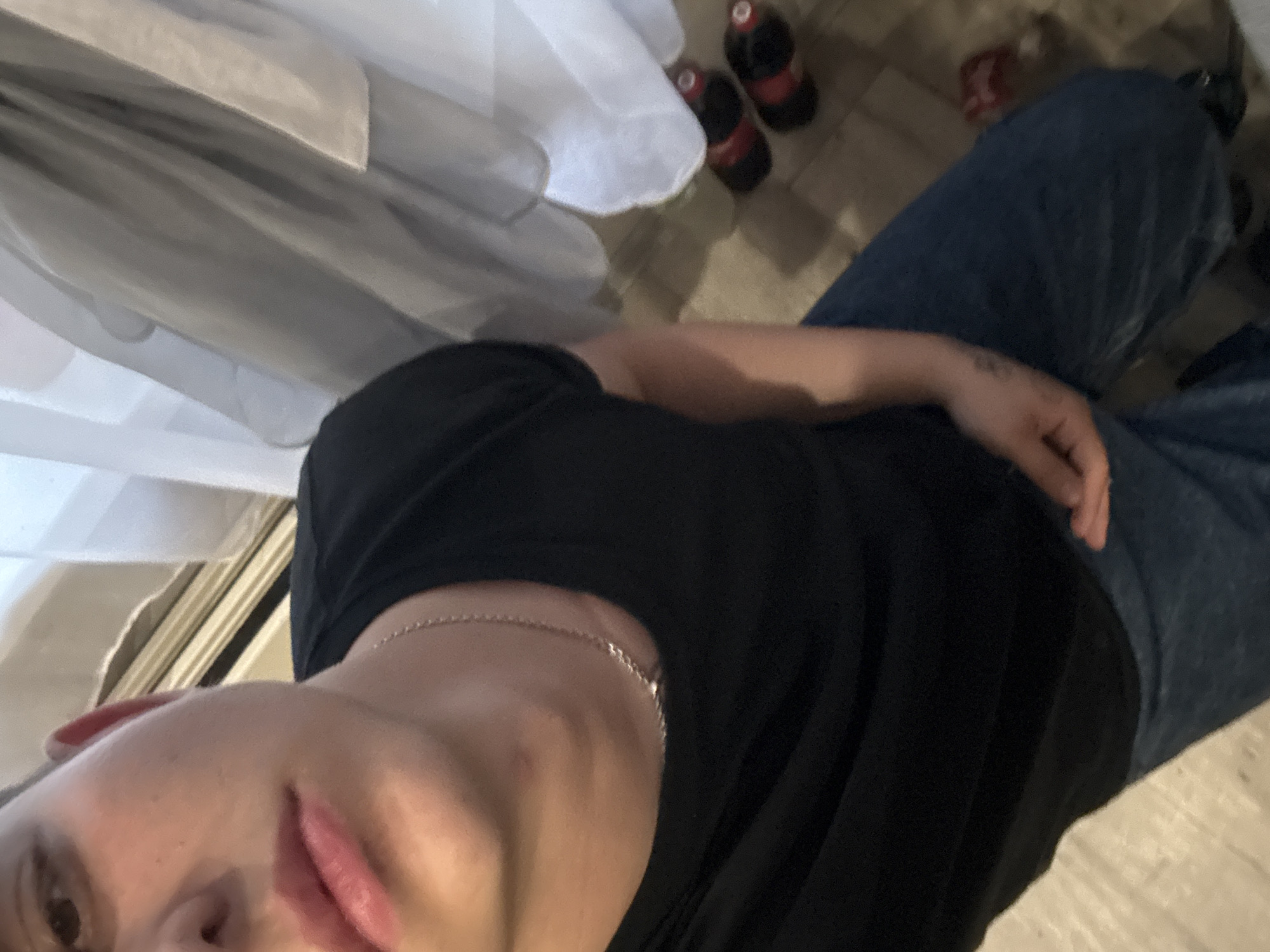 The image shows a close-up of a person from the chest up, wearing a black t-shirt and blue jeans. The person has a silver chain around their neck. Their left arm is visible, with tattoos on it. The background includes white curtains, a tiled floor, and two red and black soda bottles. The lighting is dim, casting shadows on the floor. The image is a selfie, taken from a slightly high angle. There is no information in the image to suggest it is located in Birobidzhan, Russia.