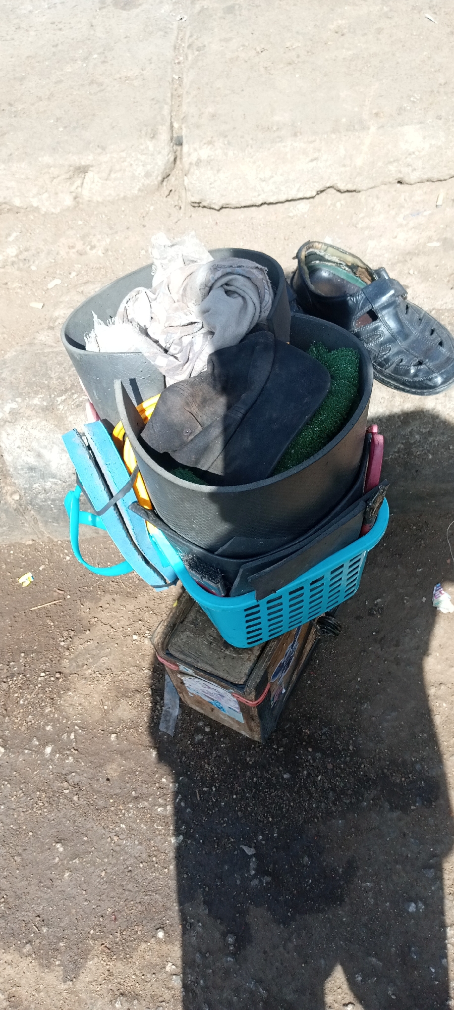 The image shows a stack of containers and objects in what appears to be an outdoor setting. A weathered, rectangular wooden box sits on the ground, topped by a light blue plastic basket. Stacked on top of the basket are two dark, round containers filled with various items, including a gray cloth, a black cap, and green material. A single black sandal rests near the top container. The ground is dirt and scattered debris. The location is likely in Gura Top, Nigeria. No people are visible.
