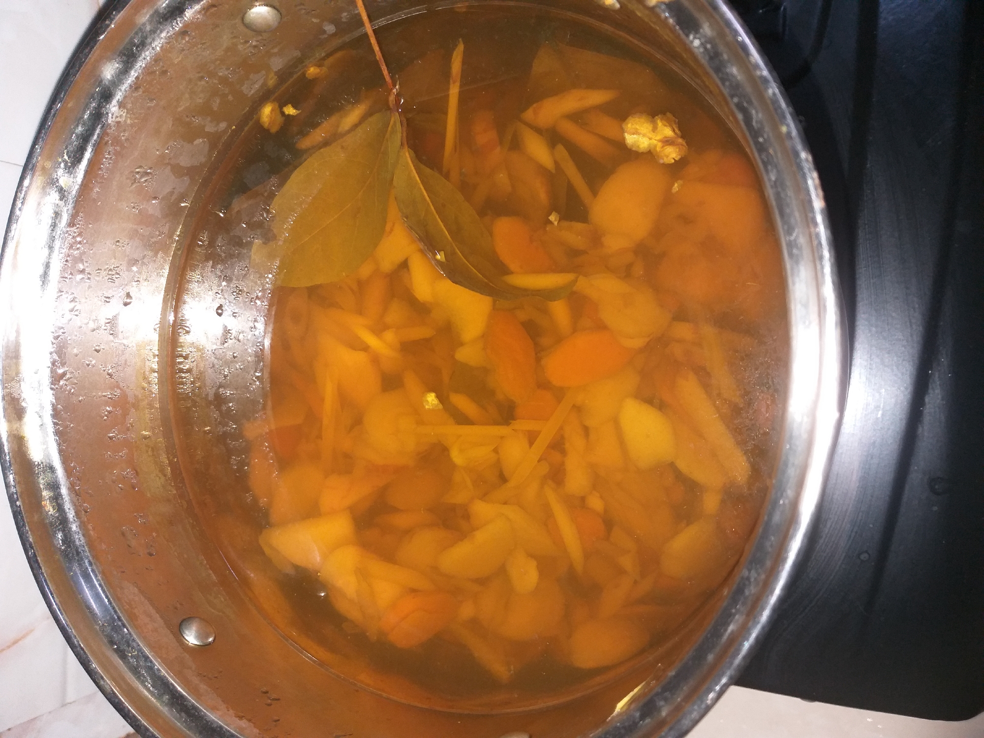 The image shows a close-up, overhead view of a stainless steel pot filled with a golden-brown liquid and various ingredients, likely being prepared on a black stovetop. The contents of the pot include sliced ginger or turmeric, and two bay leaves floating on the surface. The liquid is slightly murky, suggesting it is a decoction or soup. The pot itself appears to be a standard kitchenware item, with visible rivets on the interior. The background is out of focus, showing a patterned surface that could be a kitchen backsplash or countertop. There is no indication of location within the city of Air Jamban, Indonesia, as the image only shows a domestic cooking scene.