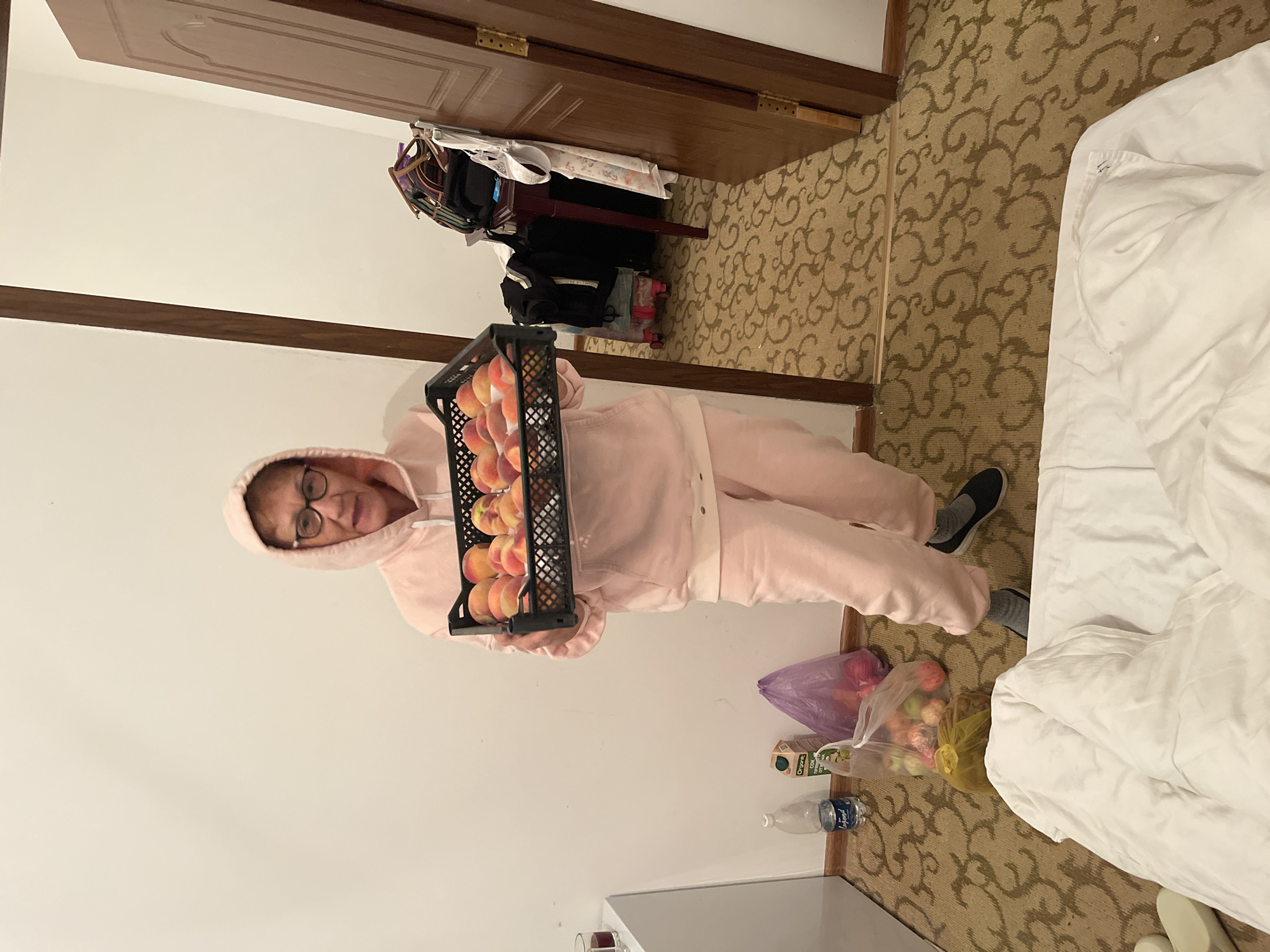 A woman wearing a pink hooded sweatshirt and sweatpants holds a black plastic crate filled with peaches. She is standing in a room with cream-colored walls and a patterned carpet. To her left, on the floor, are a plastic bottle of water, a carton of juice, and two plastic bags containing fruit. To her right, a doorway leads to another room with luggage and clothing visible. The room appears to be a hotel room. The location is the city of Ornek, Kyrgyzstan.