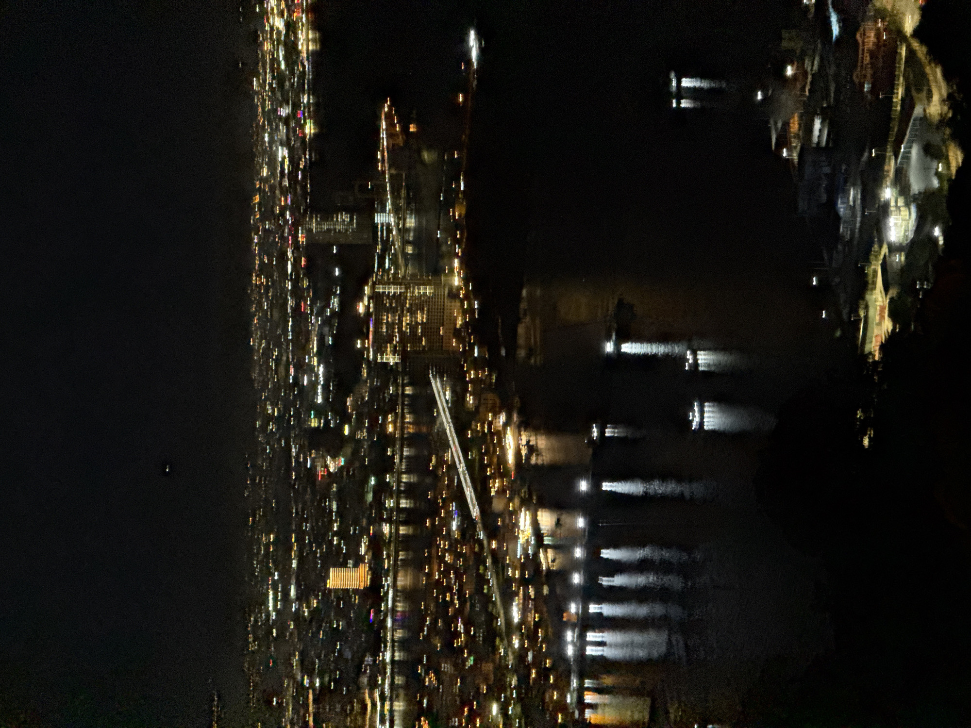 This is a high-angle nighttime aerial photograph of a city with a large body of water, likely the Han River in Đà Nẵng, Vietnam. The city lights are blurred due to motion or long exposure, creating streaks of light across the landscape and reflecting in the dark water. Several tall buildings are illuminated, including a prominent orange-lit skyscraper on the left side of the image. A long, illuminated bridge spans across the water, connecting different parts of the city. The water is dotted with the lights of moving boats, creating elongated reflections. In the foreground, the darker, undeveloped hills of the city are visible, with scattered lights indicating some structures. The sky is dark with no visible stars or moon, but a faint, small, bright object, possibly an airplane or drone, is visible in the upper portion of the frame.