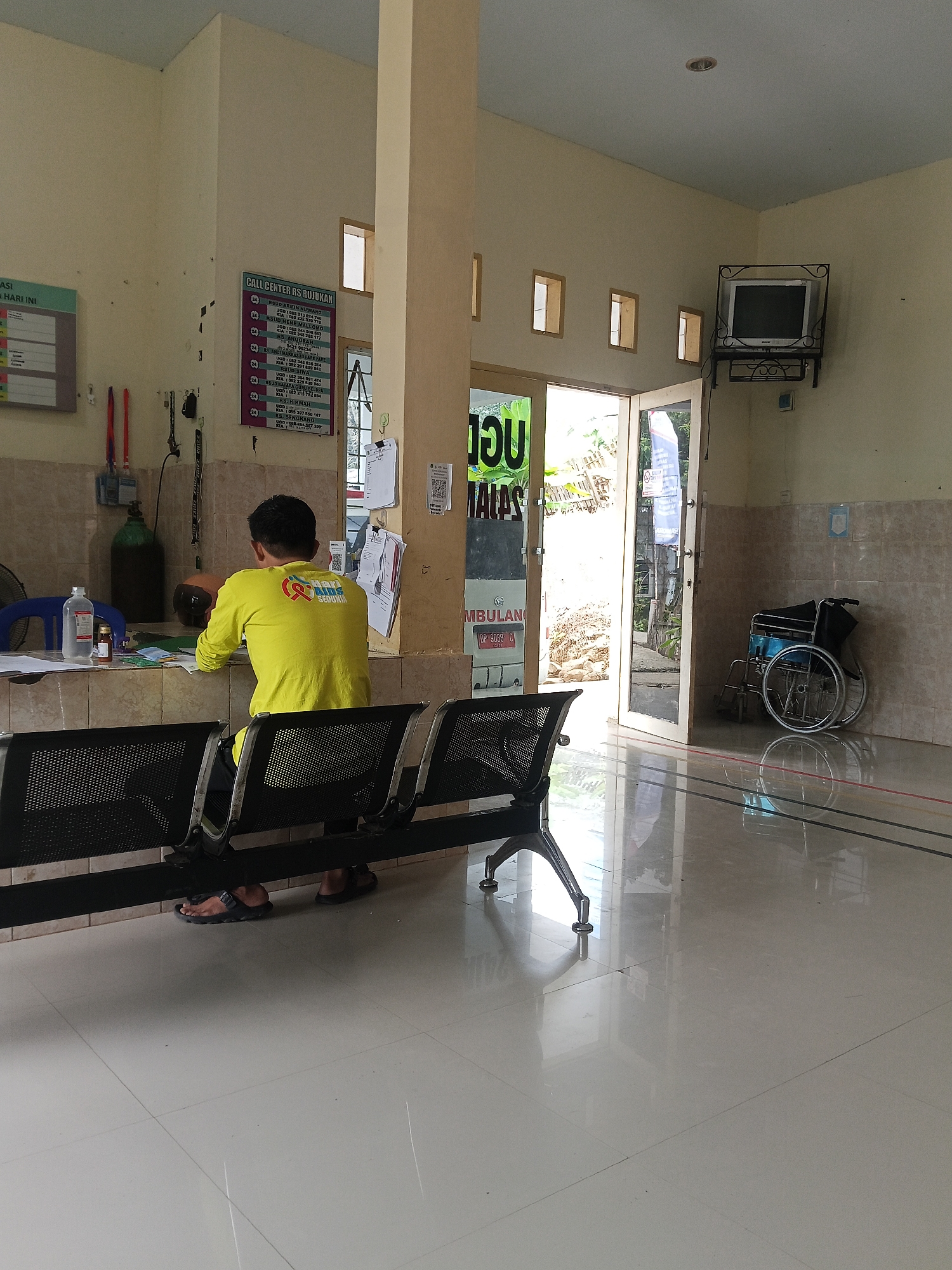 This image depicts the waiting area of a medical facility, likely a clinic or hospital, in Marannu, Indonesia. In the foreground, a man wearing a bright yellow long-sleeved shirt and dark pants sits facing away from the camera, his back to the viewer, at a reception desk. He is looking down at papers on the desk. To his left, a blue chair is visible, and on the desk are a bottle of liquid, a small brown bottle, and other documents. In the background, to the left, is a green oxygen tank. The waiting area features black metal chairs arranged in rows. To the right of the man, a doorway leads outside, revealing a glimpse of foliage. A wheelchair is parked against the wall near the doorway. Mounted on the wall above the doorway is an old television set. A bulletin board with a list of call center numbers for various hospitals is displayed on the wall to the left of the doorway. The floor is tiled and highly reflective, showing the reflections of the ceiling lights and furniture. The walls are painted a pale yellow and are partially covered with beige tiles.