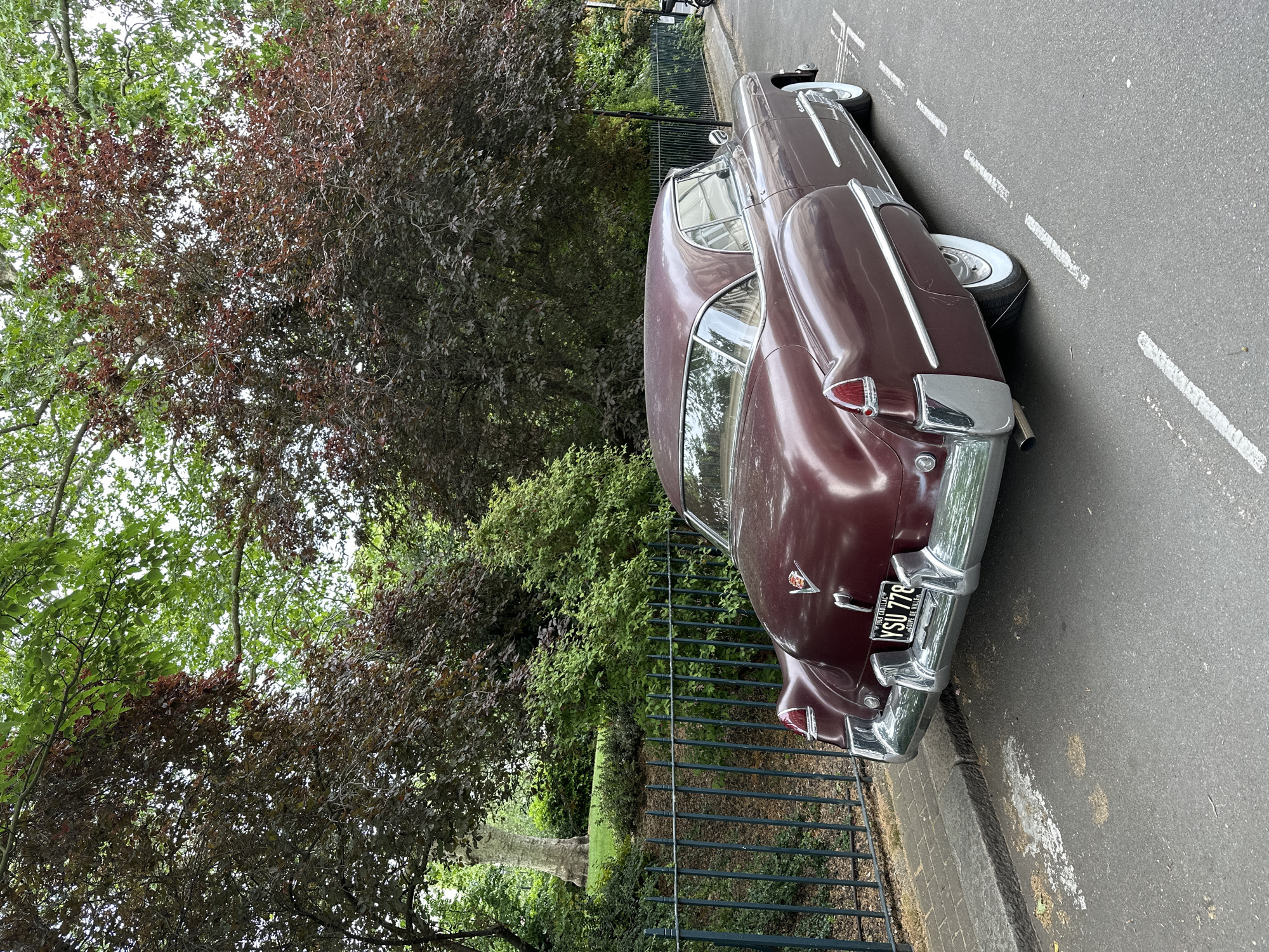 The image shows the rear of a vintage, burgundy-colored car parked on a street. The car's license plate reads "YSU 778" and indicates it is a vehicle from the US Department of the Interior. The car is situated on a gray asphalt road, adjacent to a black metal fence. Beyond the fence is green foliage, including trees and bushes. The photograph was taken in London, United Kingdom. The exact location is not clear from the image. There are no people visible in the picture. The focus is on the car and its surrounding environment.