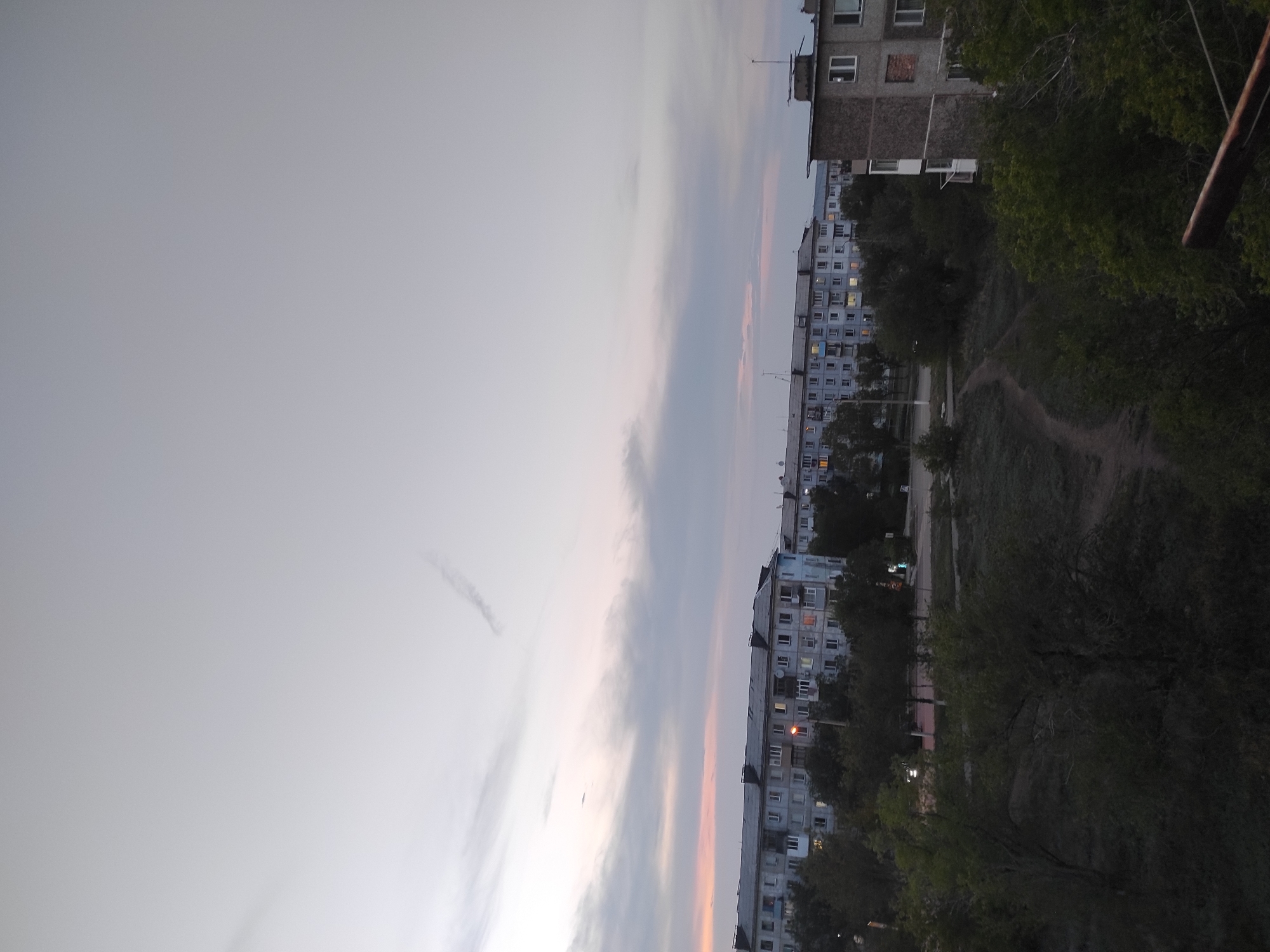 This is a vertical, eye-level, wide shot taken at dusk or dawn in Shakhtinsk, Kazakhstan. The image captures a view of several Soviet-era apartment buildings, some with lights on inside, surrounded by trees. In the foreground, there are trees and a dirt path winding through a grassy area. The sky is cloudy with hints of pink and orange from the setting or rising sun. The buildings appear to be constructed from concrete and have a consistent architectural style typical of the Soviet era. There are no people visible in the frame.