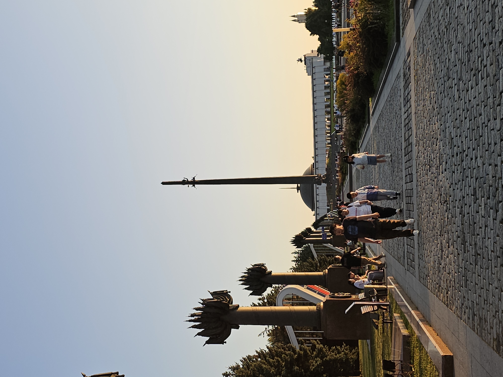 The image depicts the Victory Park on Poklonnaya Hill in Moscow, Russia, during the golden hour. A cobblestone pathway is in the foreground, leading towards a large, white, colonnaded building. To the left of the pathway, there are several tall, decorative pillars topped with stylized flames, reminiscent of eternal flames or floral motifs. Behind these pillars, evergreen trees form a dense backdrop.
In the middle ground, a tall obelisk, a prominent landmark of the park, rises against the clear, pale sky. The building behind the obelisk is long and rectangular with numerous windows or openings. A statue of a man on horseback is visible on top of this building. Further in the distance, a church with twin spires can be seen.
Several people are present in the scene. In the foreground, a man walks towards the viewer, wearing a dark t-shirt and dark pants. To his right, a young girl in a white top and skirt walks away from the viewer, and a young boy in a white and blue jacket walks beside her. Further down the pathway, more people are visible, some walking and others sitting on benches. The park appears to be a public space, with visitors enjoying the late afternoon or early evening light. The overall atmosphere is serene and pleasant, with the warm sunlight casting long shadows and illuminating the structures and foliage.