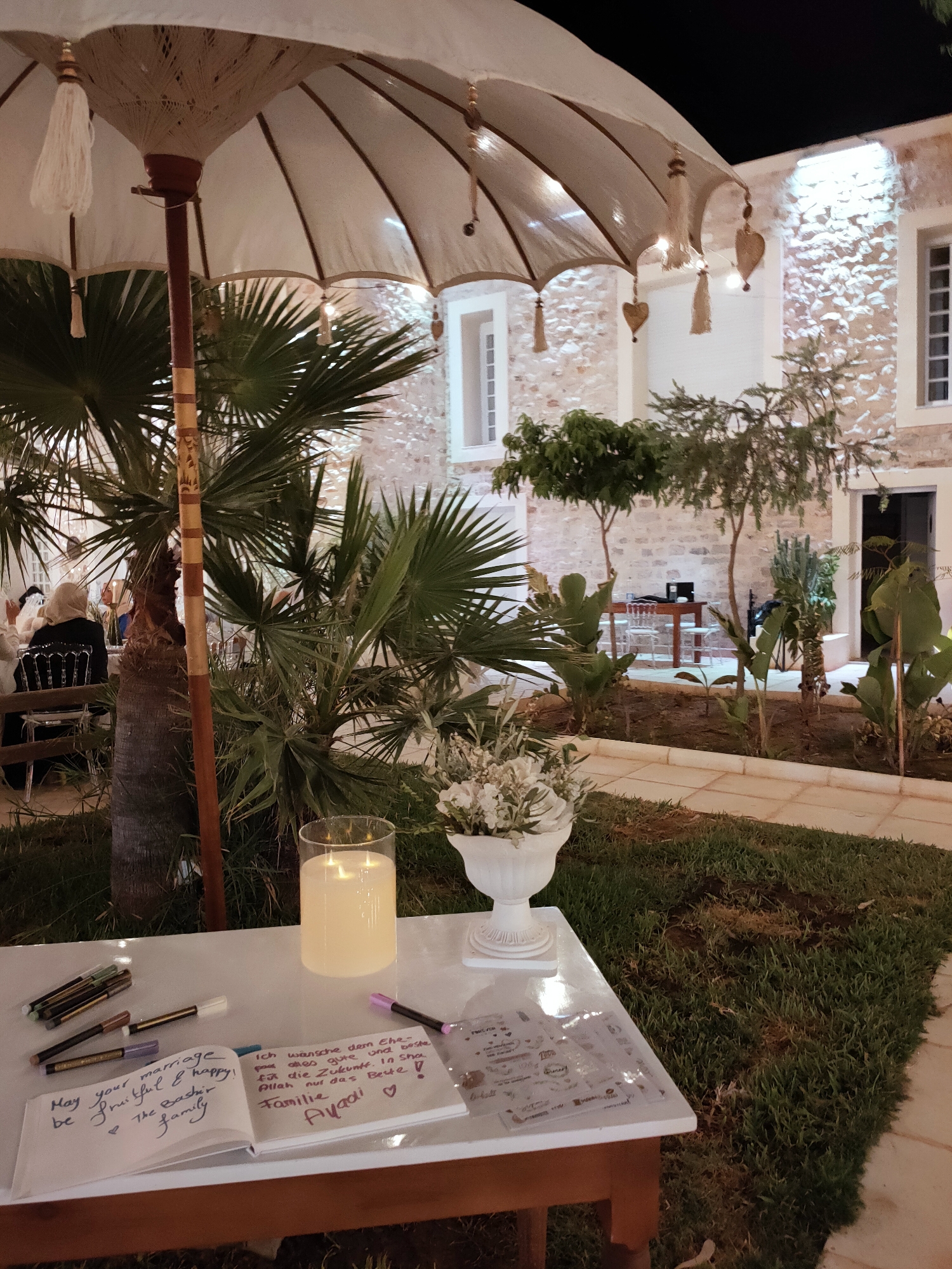 The image depicts a nighttime outdoor scene, likely a venue in Cebalat, Tunisia, decorated for an event. In the foreground, a white table is set with a large, lit cylindrical candle in a glass holder, a white vase filled with flowers, and an open guest book. A collection of pens, including several colored markers and a purple marker, are scattered around the book. The guest book has handwritten messages in both English and German. The English message reads, "May your marriage be fruitful & happy! 💖 The Backer family". The German message reads, "Ich wünsche dem Ehe-paar alles gute und beste für die Zukunft. In Sha Allah nur das Beste! Familie Ayadi ♡".
In the mid-ground, a large, ornate white umbrella with decorative tassels and heart-shaped ornaments hangs over the table and extends towards the left. Behind the table and umbrella, lush palm trees and other greenery are visible. In the background, a stone building with illuminated windows and an open doorway is lit by outdoor lighting. Several people, mostly obscured by the foliage, are seated at tables in the distance, suggesting a gathering or reception. The ground is covered with grass and a paved walkway.
