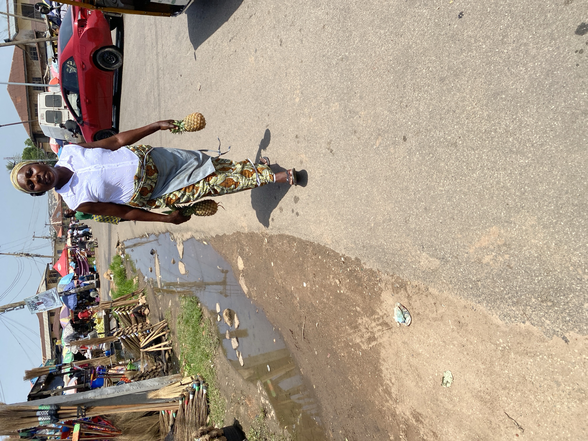 A woman is standing on a paved road, holding two pineapples. She is wearing a white shirt, a patterned skirt, and a headscarf. The road is cracked and appears to be in a market area, with stalls and vendors visible in the background. There are also puddles of water on the ground, suggesting it may have recently rained. The scene appears to be outdoors during the day, with bright sunlight casting shadows. The woman's expression is neutral. Various goods are displayed at the market stalls, including brooms and what appear to be fruits or vegetables. A red car is parked nearby. The overall setting suggests a busy outdoor market in a developing urban area. There is a banner visible in the background with some text, but it is too blurry to read.