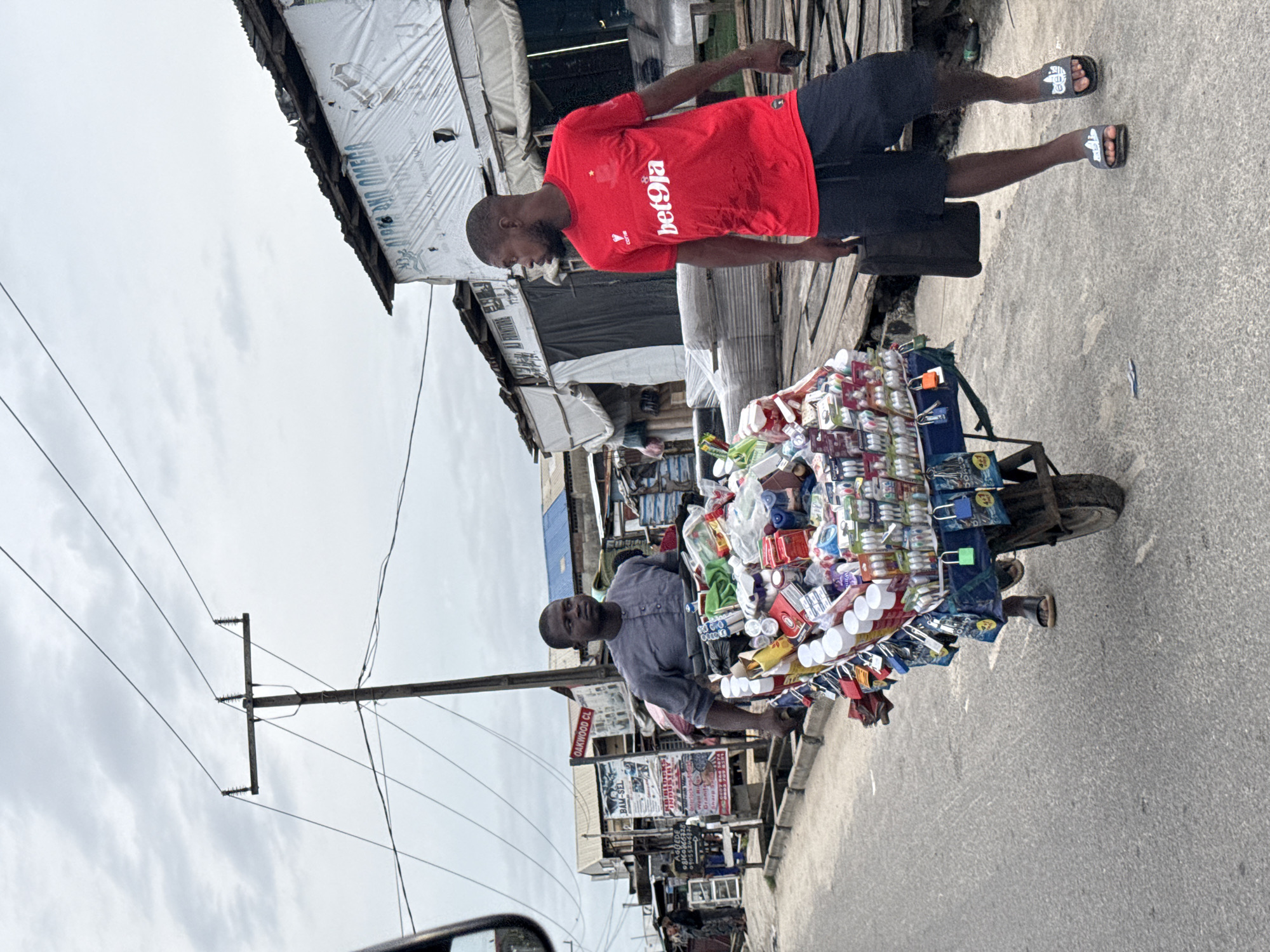 This is a street-level, eye-level, medium shot taken outdoors in Ado, Nigeria, on a cloudy day. The scene depicts a busy commercial street with various stalls and structures on either side of the asphalt road. The foreground features a street vendor pushing a cart loaded with an assortment of goods.
The cart is filled with various items, including bottled products, packaged goods, and possibly toiletries or small electronics. The vendor, a man wearing a grey shirt and dark trousers, is actively pushing the cart. To his right, another man, also a street vendor, walks alongside the cart, carrying a dark bag in his left hand and wearing a red t-shirt with "bet9ja" logo and dark shorts. He is wearing grey slides on his feet.
The background reveals a dense urban environment characterized by temporary structures and businesses. Signs with lettering, some in English, are visible on the buildings. Power lines and a utility pole suggest the presence of electricity infrastructure. The general atmosphere appears to be one of commerce and daily life in a Nigerian city.