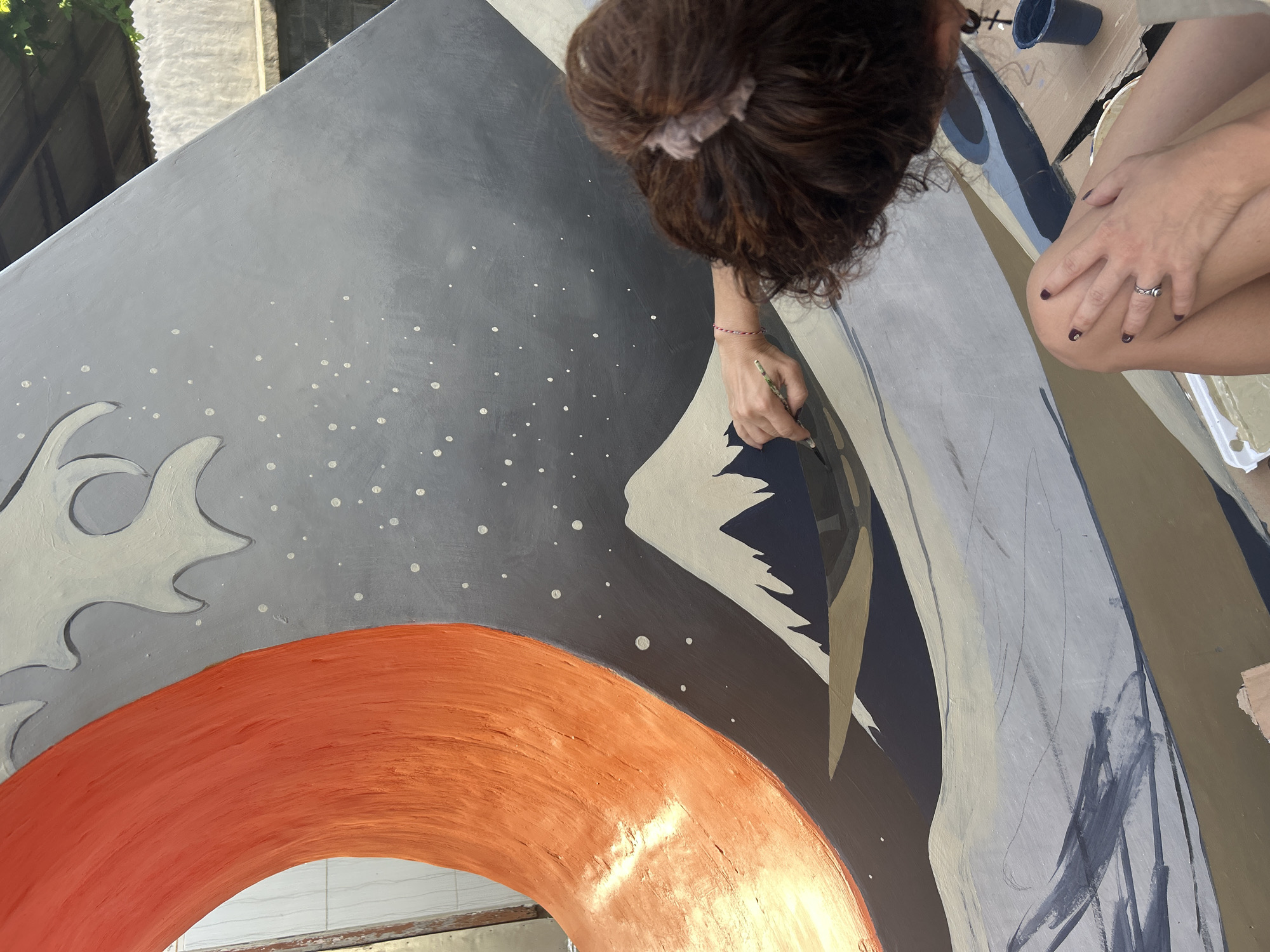 A woman with her hair in a bun is painting a mural in Kediri, Indonesia. The mural features a mostly grey-colored background with white dots, as well as an orange ring shape on the bottom left. The painting is still in progress, with some areas in grey underpainting. The woman is holding a paint brush and is working on a section of the mural. A container of blue paint is present. She is wearing a ring and has dark painted fingernails. The mural appears to be outdoors, possibly on a wall or a large panel.