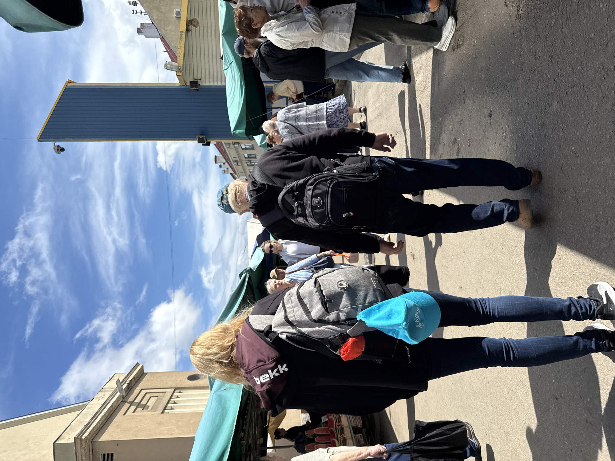 This is a candid street-level photograph taken from a slightly elevated perspective, looking down a paved walkway at an outdoor market. The lighting suggests it is daytime with bright sunlight casting distinct shadows from the people and market stalls. The market appears to be in an urban setting, likely in Riga, Latvia, given the architectural style of the buildings visible in the background which include a distinct, tall, blue structure with a yellow-trimmed roof and a more traditional light-colored building with a stylized geometric design. Green market stall awnings are prominent, creating a canopy over parts of the walkway.
In the foreground, two individuals are walking away from the camera. The person on the left has blonde hair and is wearing a dark hooded jacket with the word "bekk" visible in white lettering on the hood. They are carrying a grey backpack with a red object and a bright blue cap attached. They are wearing dark blue jeans and black and white sneakers. To their right, a man wearing a blue baseball cap is walking away, also with a black backpack. He is wearing a black jacket and dark pants.
Further down the walkway, a crowd of people are browsing or walking. One woman in a patterned dress and cardigan is visible, as is another woman in a light-colored jacket and trousers. There are market stalls with goods, including watermelons displayed on the left. The overall atmosphere is that of a busy, public marketplace on a sunny day.
