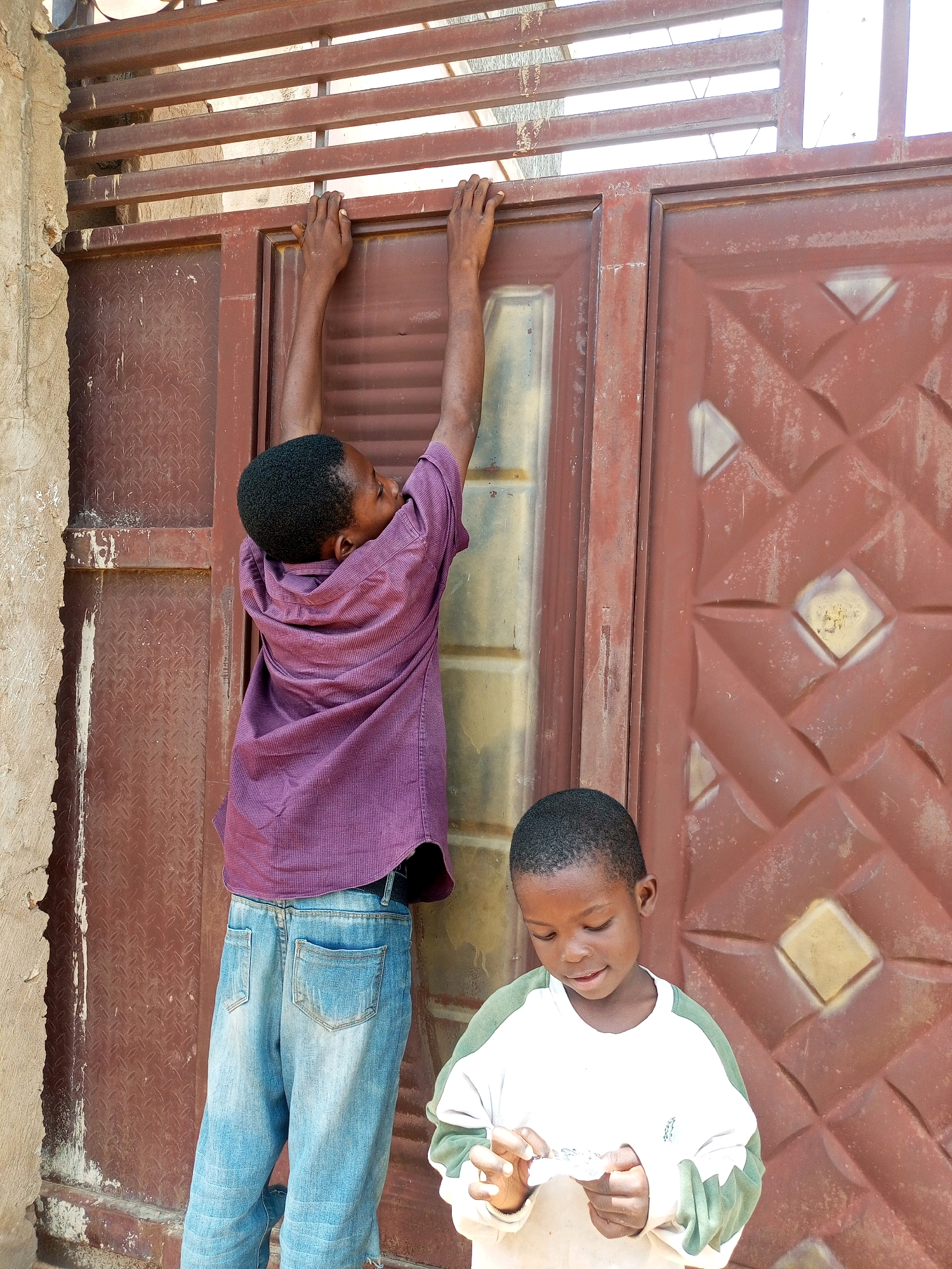 In Tempure, Nigeria, two young boys stand before a weathered, rusty metal gate. The boy on the left, wearing a purple, collared shirt and blue jeans, is reaching up with both hands to grasp the top horizontal bar of the gate. He is facing away from the camera, with his head tilted back and looking upward. His body is positioned in a dynamic pose, suggesting he is either trying to climb or reach something. To his right and slightly in front, a younger boy, wearing a white long-sleeved shirt with green accents on the sleeves and collar, looks down at his hands. He is holding a small, white object, possibly a piece of paper, and appears to be examining it or unfolding it. The gate itself is made of metal panels with a diamond-shaped embossed pattern. The surrounding environment appears to be outdoors, with a textured, light-colored wall visible on the far left. The lighting suggests it is daytime.