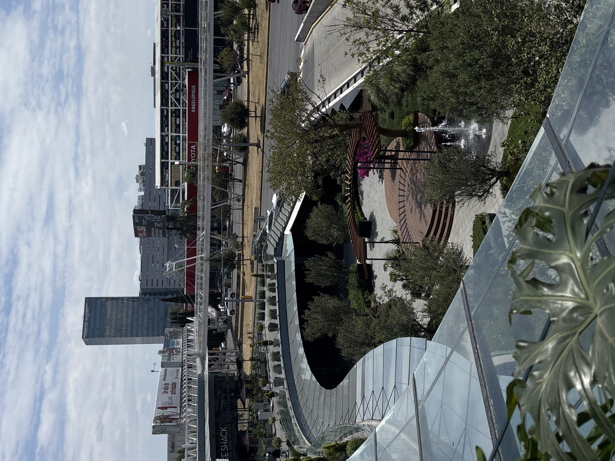 The image shows a view from a high vantage point, likely a building, overlooking a section of San Bernardino Tlaxcalancingo, Mexico.
In the distance are buildings, including a tall skyscraper labeled "Hospital Angeles," and a structure with "ANGELOPOLIS" and "YOTA" signage. There's also a "Shake Shack" visible.
The foreground features modern architectural elements with glass and curved lines. There is an open-air space that has trees, a garden with a fountain, and a pergola-like structure. A road with parked vehicles is visible beyond.
