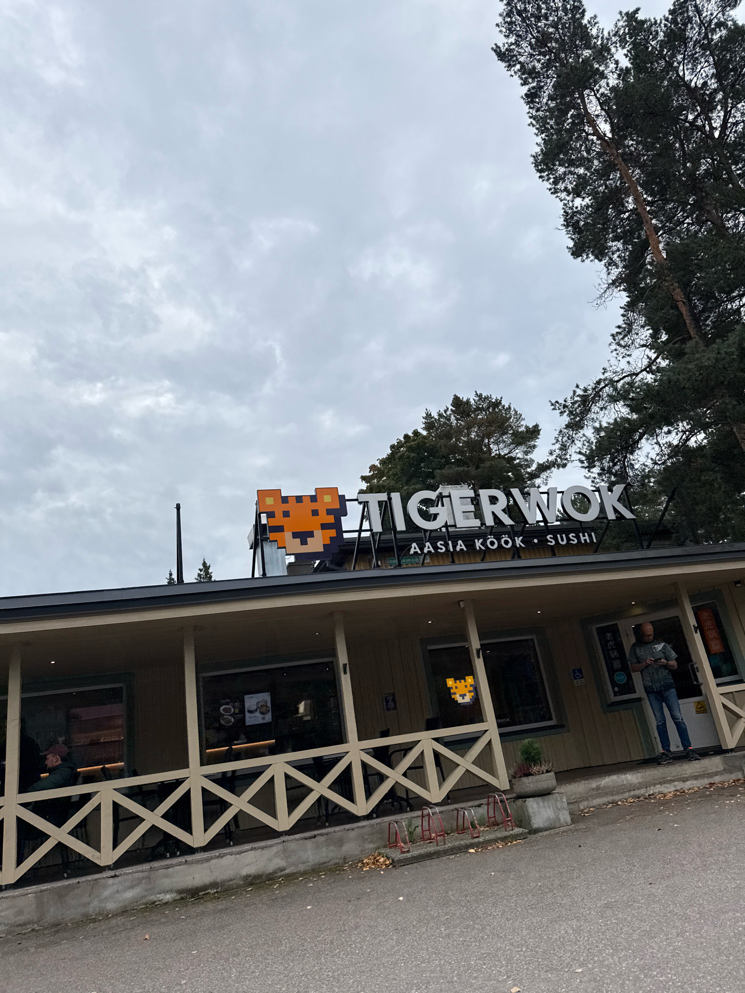 The image shows the exterior of a restaurant named "TIGERWOK" located in Tallinn, Estonia. The restaurant's sign features a pixelated tiger face and the words "TIGERWOK" in large white letters, with smaller text below reading "AASIA KÖÖK • SUSHI". The building has a light-colored facade with a covered porch area supported by wooden posts. A white wooden railing with a diamond pattern encloses the porch. There are large windows on the building, some of which display menus or interior views.
One person, a man wearing a gray t-shirt and blue jeans, is standing near the entrance of the restaurant, looking at his phone. Another person, wearing a dark jacket and a cap, is seated at a table on the porch. A bicycle rack is visible in front of the building. Trees and a cloudy sky form the background. The overall scene suggests a somewhat overcast day.