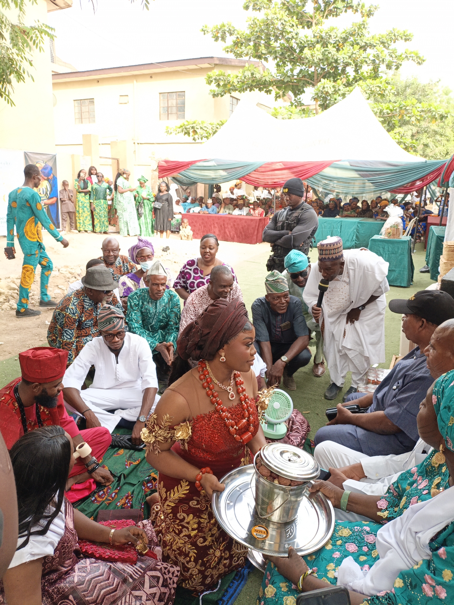 This image depicts a vibrant outdoor gathering, likely a traditional Nigerian ceremony or celebration, taking place in Sabon Gari West, Nigeria. The scene is bright and sunny, filled with numerous people dressed in colorful, elaborate traditional attire.
In the foreground, a central figure, a woman adorned in a rich red and gold embroidered dress, a matching headwrap (gele), and a prominent red beaded necklace, sits on the ground. She holds a large silver tray containing a metal bucket filled with nuts, possibly kola nuts, which are significant in Nigerian cultural rituals. A small green portable fan sits on the ground near her, hinting at warm weather. Around her, several men and women are seated on green matting, engaged in conversation or observing the proceedings. Notable figures include a man in a red agbada and cap holding a pipe, and a woman in a teal and gold floral dress to the right of the tray.
In the mid-ground, a man in a white agbada and patterned cap addresses the attendees with a microphone, suggesting a formal part of the event. Nearby, a man in a black tactical vest and beanie stands guard. Guests are seated under white tents draped with red and green fabric, which are consistent with Nigerian event decor. To the far left, a man in a distinctive light blue and yellow patterned jumpsuit walks past a banner with a partial image of a person in traditional clothing and text that includes "SPORTS" and "YOUTH".
The background features a light-colored building and lush green trees under a clear sky. The overall atmosphere is festive and communal, with a sense of respectful gathering. Visible text on the silver tray includes "NOVA" and "40CM".
