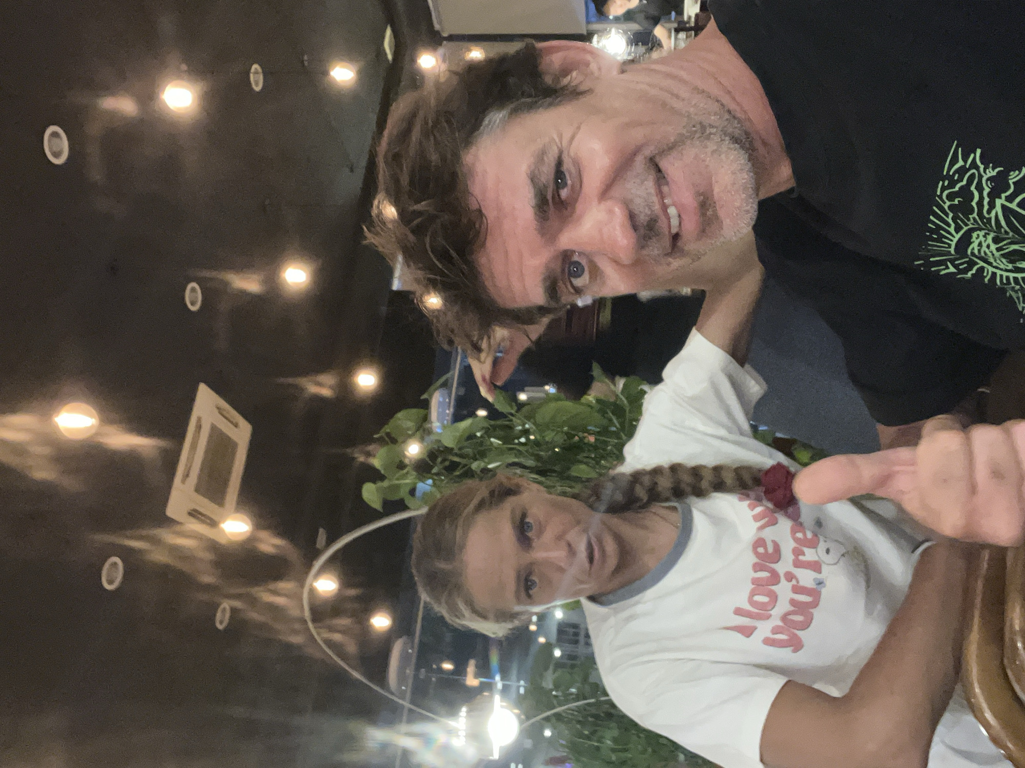 In Phu Thuy Ward, Vietnam, a selfie is taken indoors, likely in a restaurant, as indicated by the overhead lighting and ceiling-mounted air conditioning unit. The image captures two individuals: a woman with blonde hair in a braid wearing a white t-shirt with the text "I love you're", and a man with brown hair and a beard giving a thumbs-up. The lighting suggests it is evening or nighttime, with warm yellow lights illuminating the scene. The background is somewhat blurred, but a plant is visible to the left of the woman, and other patrons and possibly a bar area can be glimpsed in the distance. The overall atmosphere appears casual.