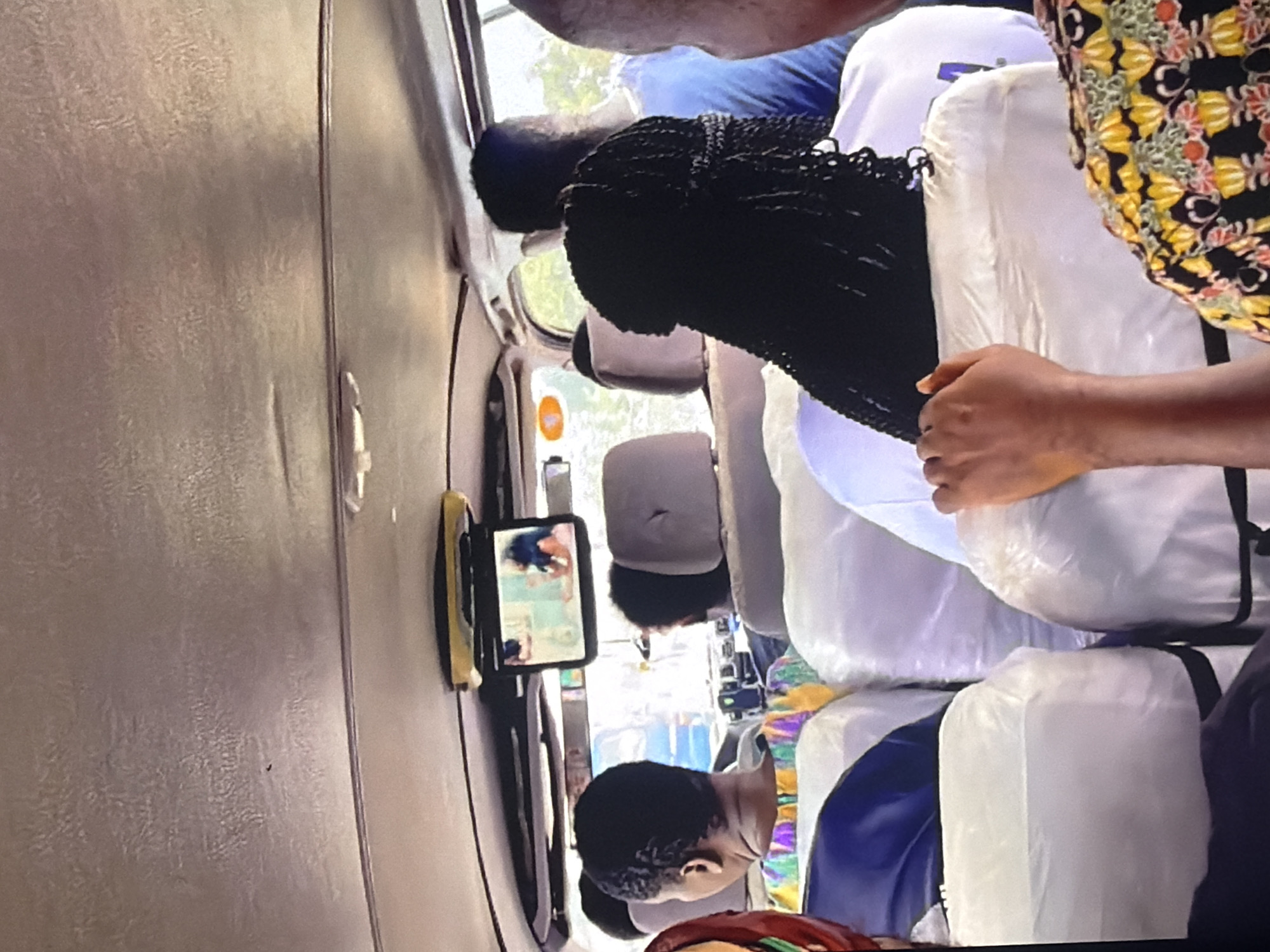 The image depicts the interior of a public transport vehicle, likely a minibus or van, on a street in Bukuru, Nigeria. The ceiling of the vehicle is visible, showing a textured, light brown material. A small screen is mounted to the ceiling, displaying a video of two women. Below the screen, several passengers are seated, their heads and shoulders visible. The seats appear to be covered in white plastic. One passenger in the foreground has dark, braided hair and is wearing a white garment. Another passenger to their left has short, dark hair and is wearing a colorful, patterned shirt. Outside the vehicle, trees and greenery are visible through the windows, suggesting an outdoor setting in a well-vegetated area of Bukuru. The lighting indicates it is daytime.
