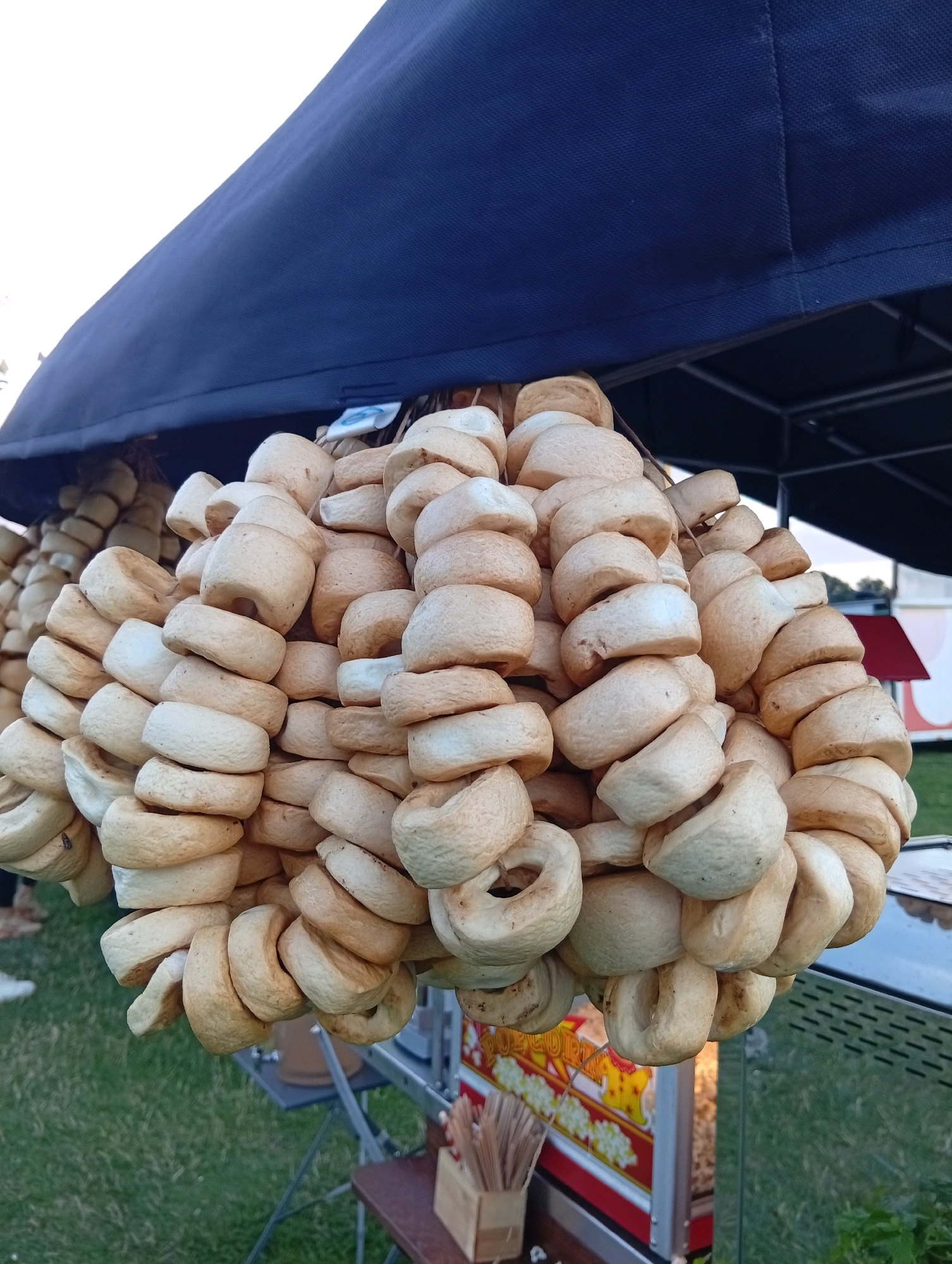 A close-up, low-angle shot captures strings of baked goods, resembling bagels or ring-shaped bread, hanging from a dark blue canopy. The baked goods are light brown and appear to be arranged in multiple strands. In the background, a popcorn machine is visible, with a bright red and yellow graphic. Beyond that, a green grassy area and a small building with a red roof can be seen. The scene is likely an outdoor market or fair, possibly in Szewce, Poland, given the context. The lighting suggests it is daytime, with a bright sky visible in the upper left corner. There are no people clearly visible in the frame, although the suggestion of a person's leg can be seen on the far left.