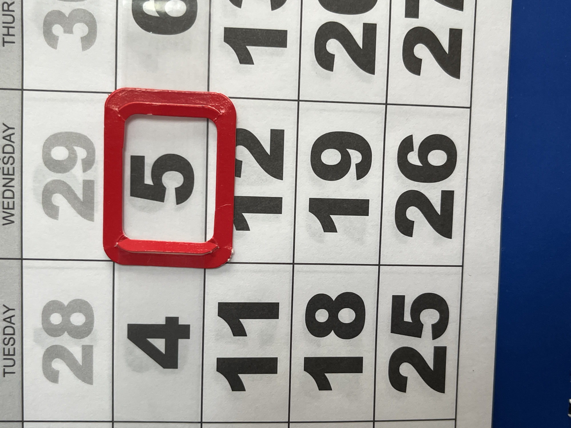 A close-up, overhead shot shows a grid calendar. The days of the week are printed at the top: TUESDAY, WEDNESDAY, THURSDAY. Numbers are printed within each cell of the calendar grid. The number 28 is in the TUESDAY column, and 29 is in the WEDNESDAY column. A bright red, rectangular marker is placed over the number 5 in the WEDNESDAY column, which is in the second row from the top. Below the 28 and 29, the numbers 4, 5, and 6 are visible in their respective columns. Further down, the numbers 11, 12, 13, 18, 19, 20, 25, 26, and 27 are visible. The bottom of the image shows a blue surface. This image does not contain any information that would allow it to be placed in Saratov, Russia.