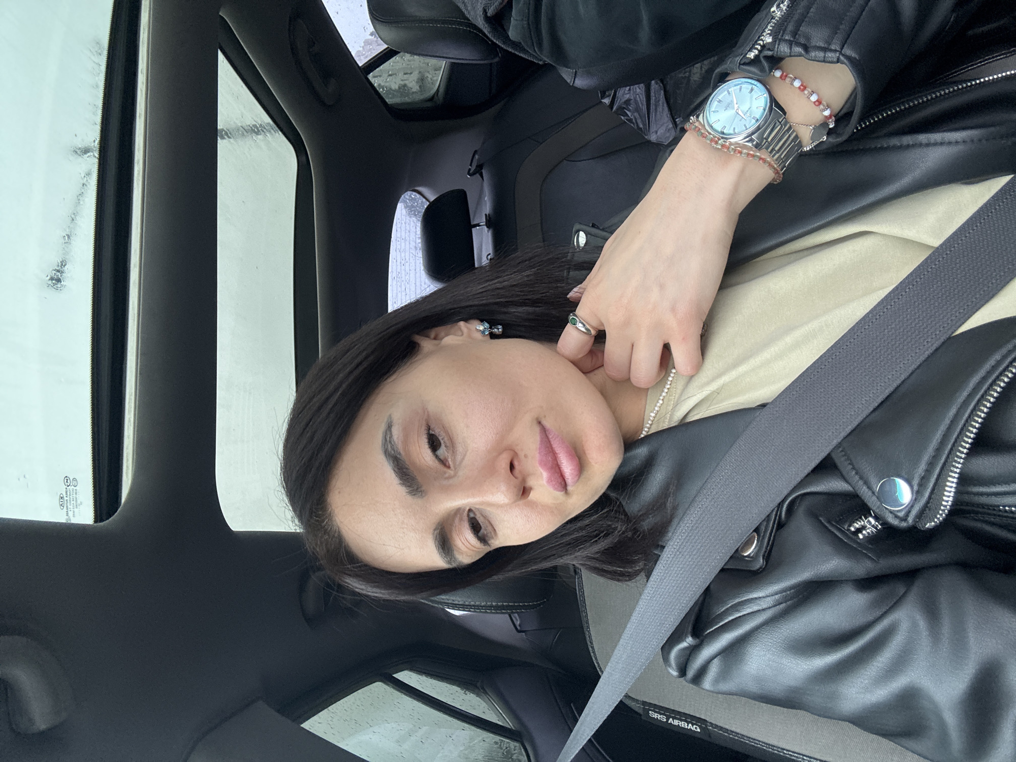 A forensic examination of the image reveals an adult female seated in the passenger side of a dark-interior vehicle, likely a KIA based on faint mirrored text visible on the front windshield. She is secured by a grey shoulder-and-lap seatbelt, which crosses her black faux leather jacket and a light beige top. The jacket features silver zippers and snap buttons. Her dark, chin-length hair frames her face, which exhibits light makeup, including pink lipstick. She wears a white beaded necklace, a blue multi-stone earring in her left ear, a silver ring with a green stone on her left index finger (with her left hand positioned near her neck), a silver watch with a light blue face, and two beaded bracelets (one red/orange, one white/pink) on her right wrist. Her gaze is directed towards the camera.
The vehicle interior features dark-colored leather seats and dark grey trim. An "SRS AIRBAG" label is visible on the grey panel next to the passenger seat. Above, a panoramic sunroof is present, showing a wet, overcast sky, indicating rain. The rear seat area is dark, with an indistinct figure possibly present in the far right passenger seat. Through the rear window and sunroof, the exterior appears rainy with blurred background elements suggesting an urban or suburban setting.
While the scene is within a vehicle during rainy conditions, no specific landmarks, street signs, or unique architectural features are discernible within the image to pinpoint an exact location within the city of Tbilisi, Georgia.